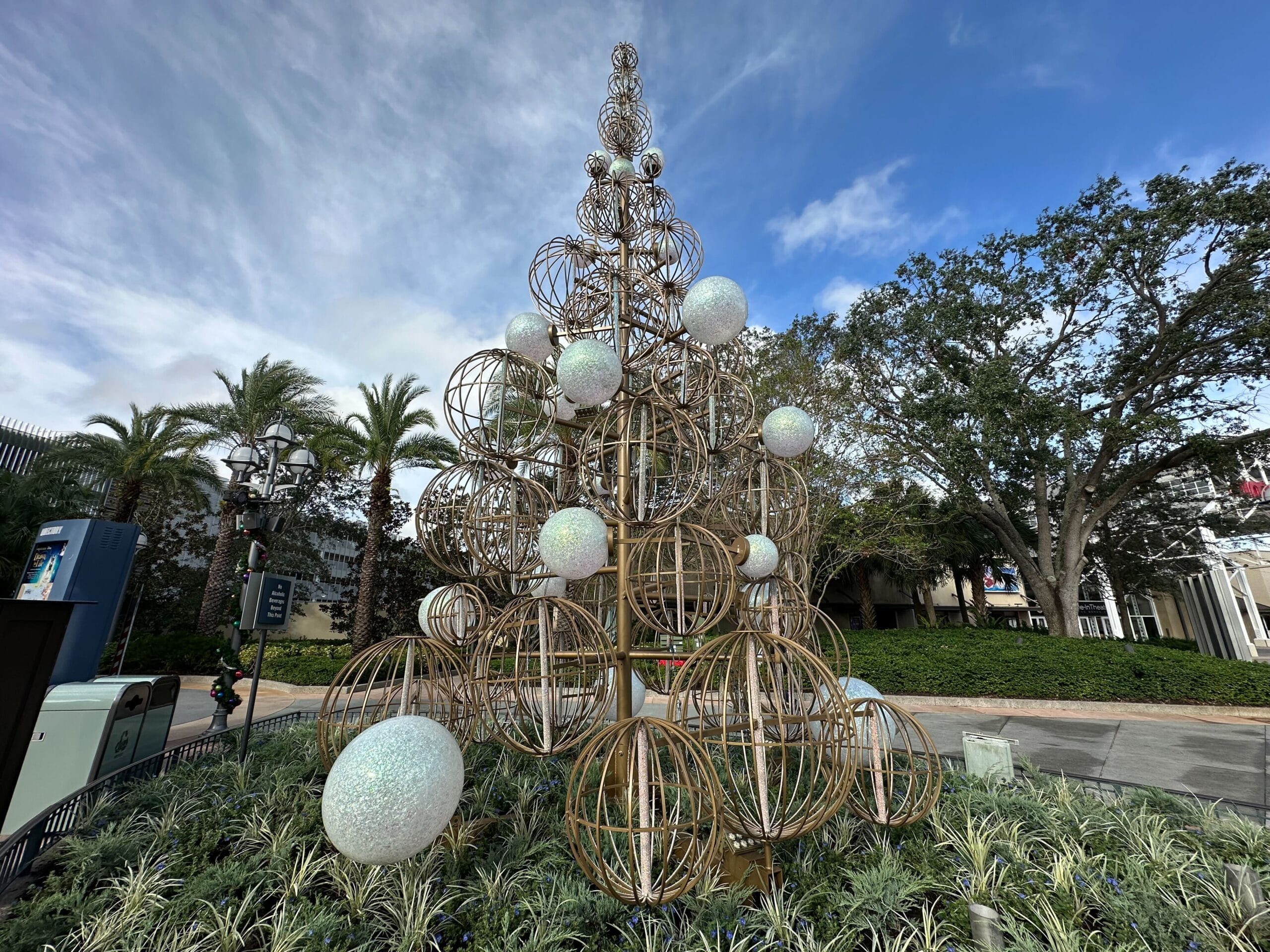 Large sculpture of a Christmas tree made of metal spheres and ornaments is displayed outdoors, surrounded by greenery and palm trees.