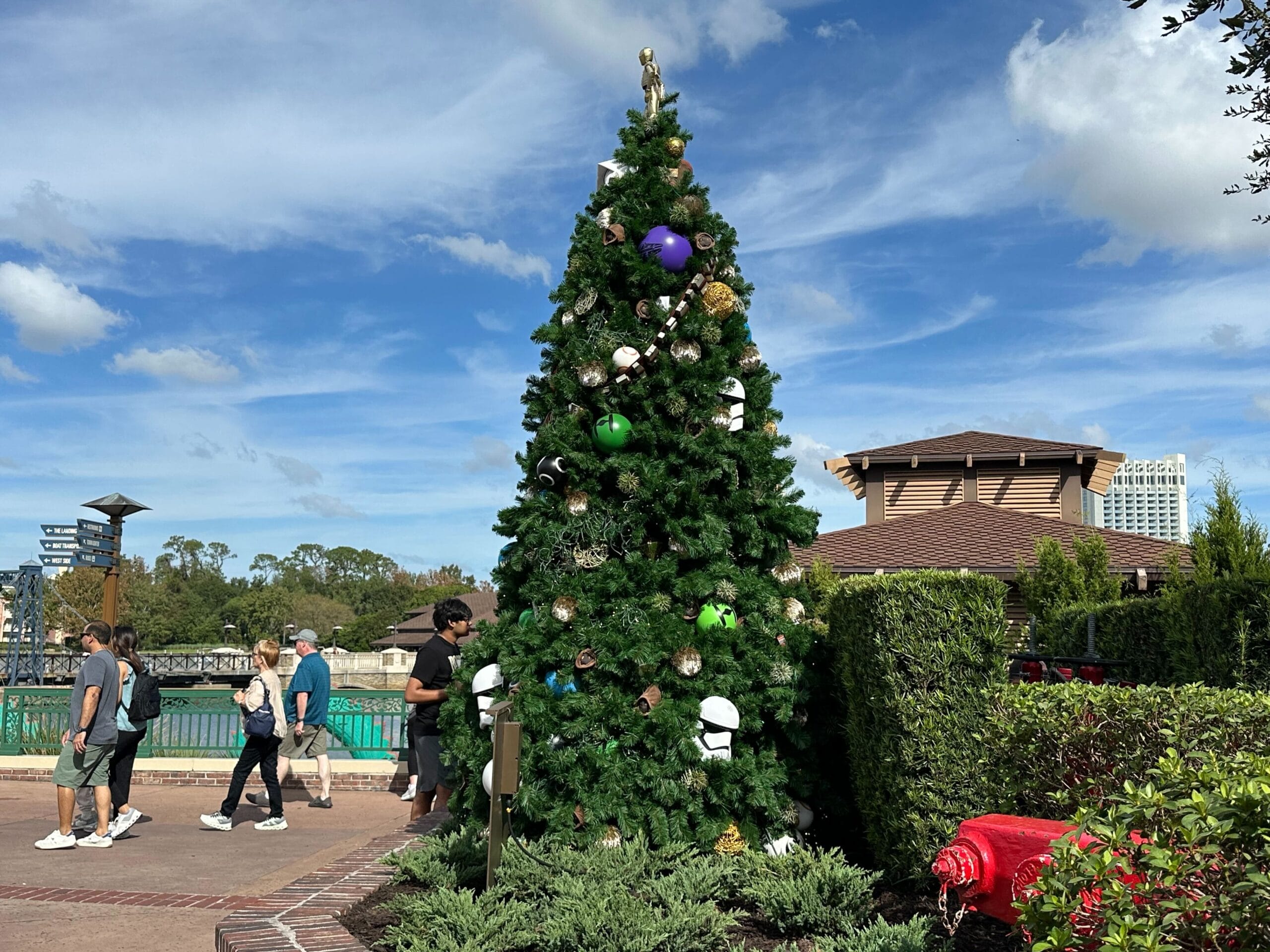 A large Christmas tree decorated with themed ornaments stands outdoors near a paved pathway. People walk by under a clear blue sky with scattered clouds.