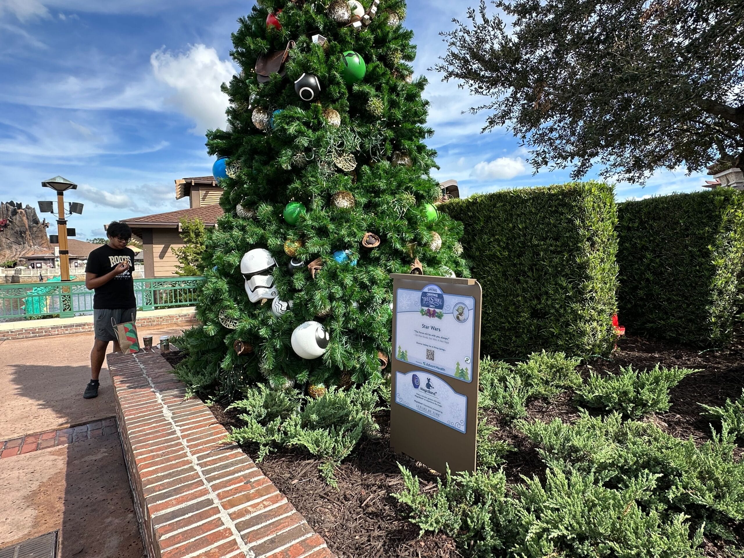 A Christmas tree decorated with Star Wars helmets and ornaments stands by a sign next to a walkway, with a man walking nearby.