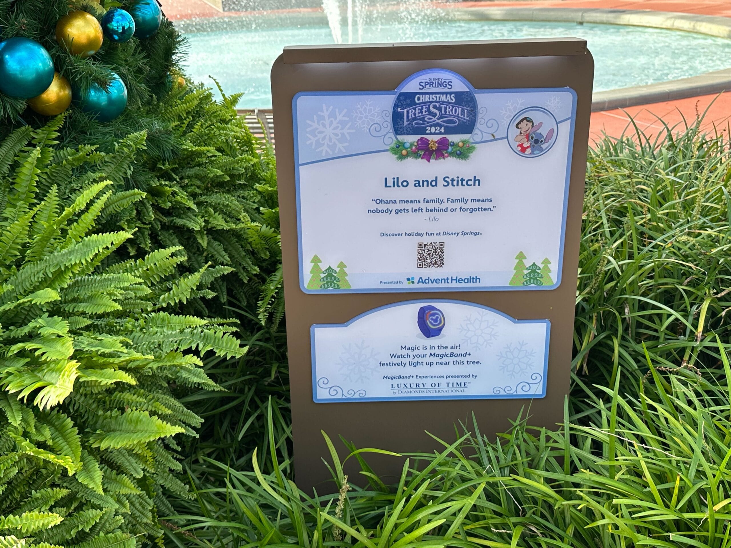 A holiday-themed sign for "Lilo and Stitch" at Disney Springs, surrounded by grass and part of a decorated tree visible on the left.