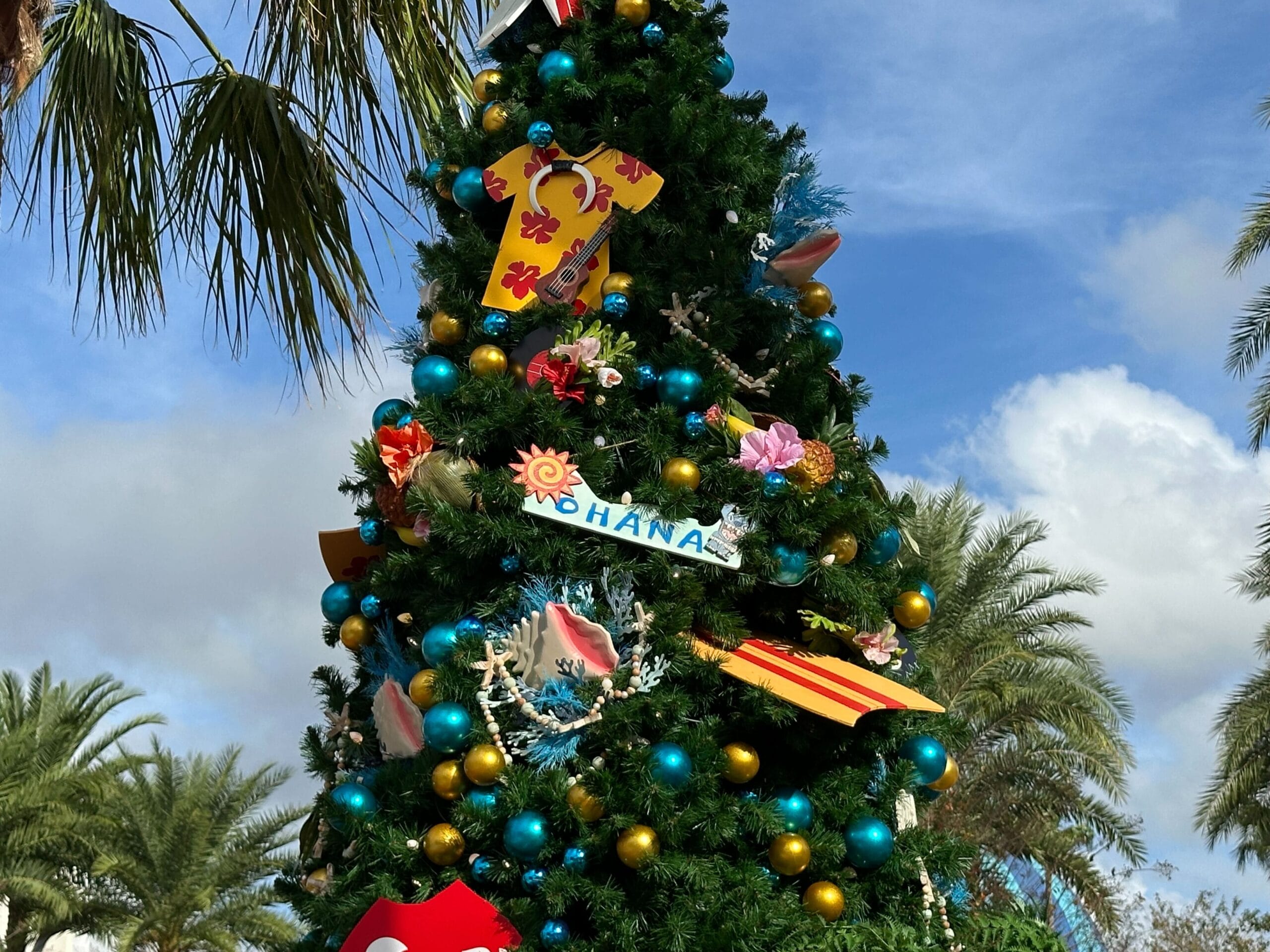 Decorated Christmas tree outdoors with tropical-themed ornaments and blue and gold baubles, surrounded by palm trees under a cloudy sky.