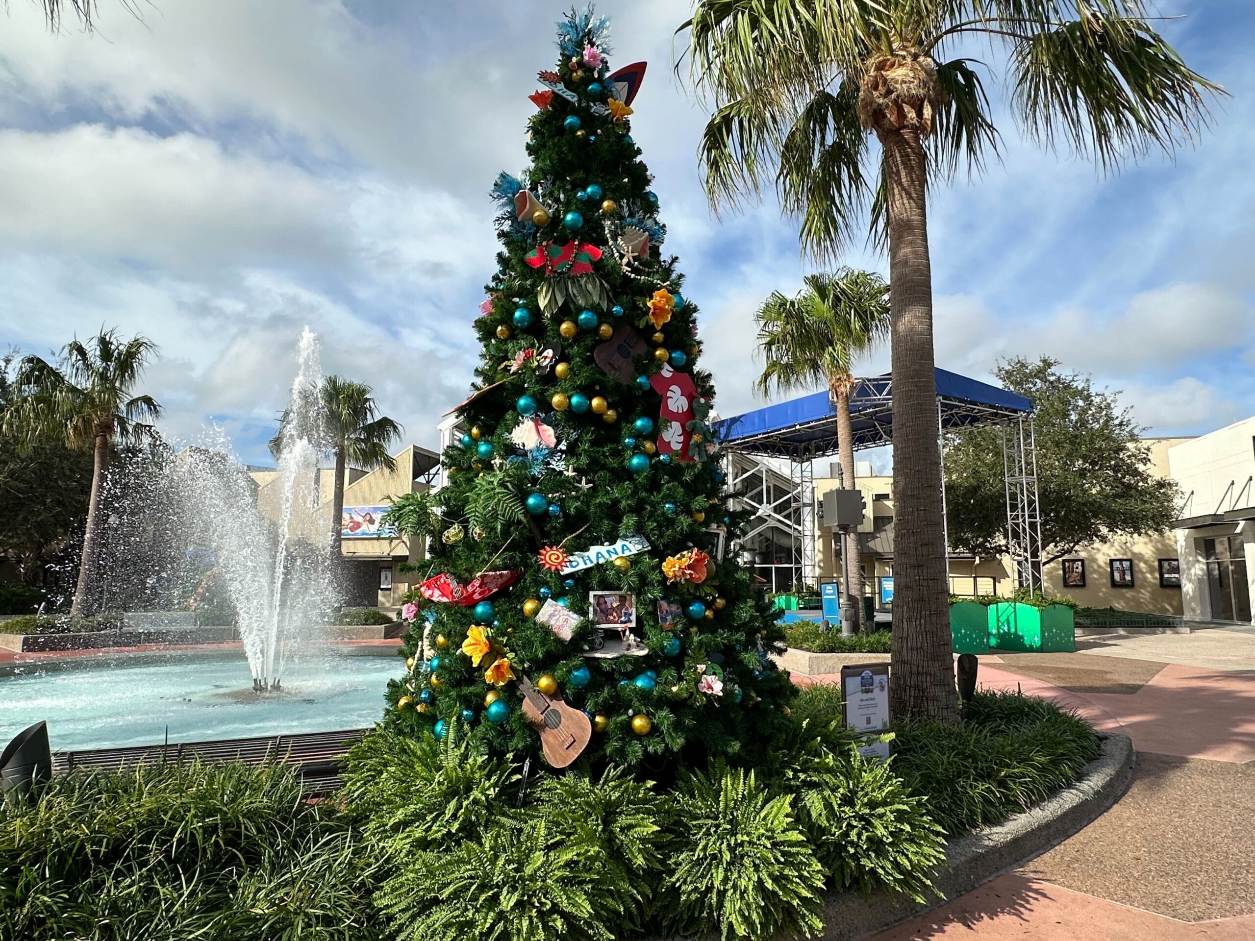 A decorated Christmas tree stands outdoors near a fountain, surrounded by palm trees and grassy plants under a partly cloudy sky.