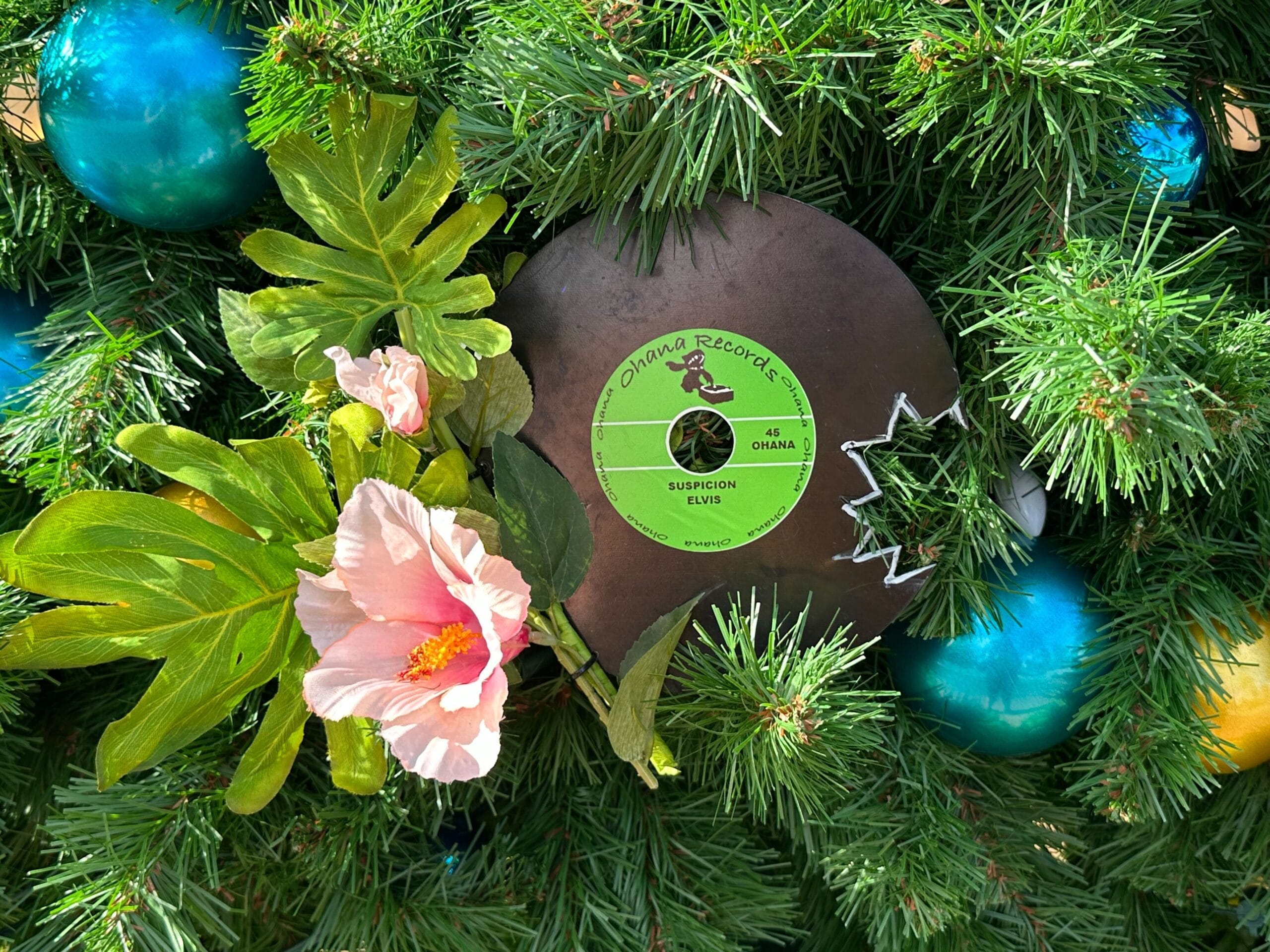 A vinyl record with a green label is nestled in a Christmas wreath with pink flowers and green ornaments.