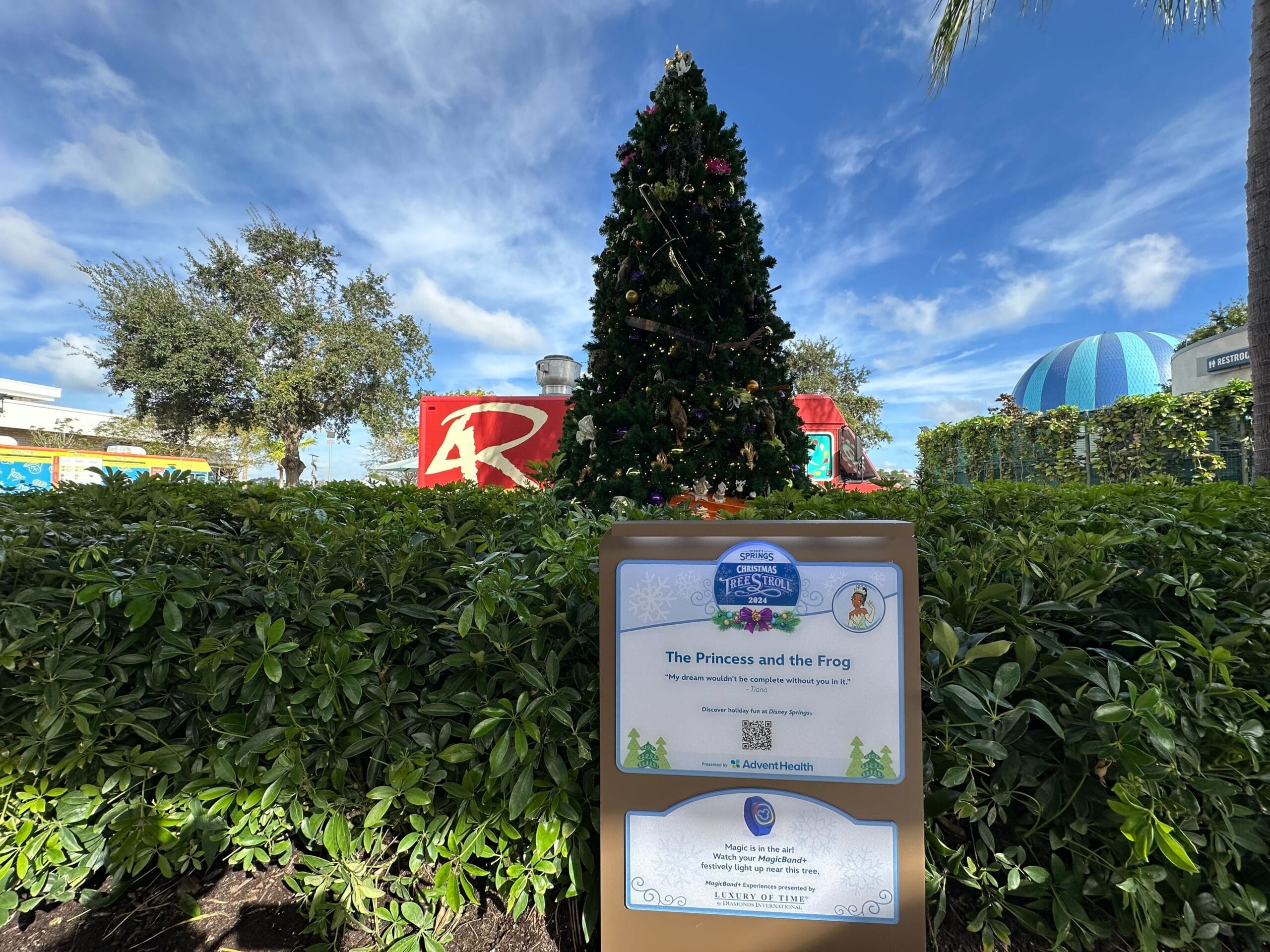 Decorated Christmas tree surrounded by bushes and greenery, with a sign reading "The Princess and the Frog" in front of a themed backdrop under a blue sky.
