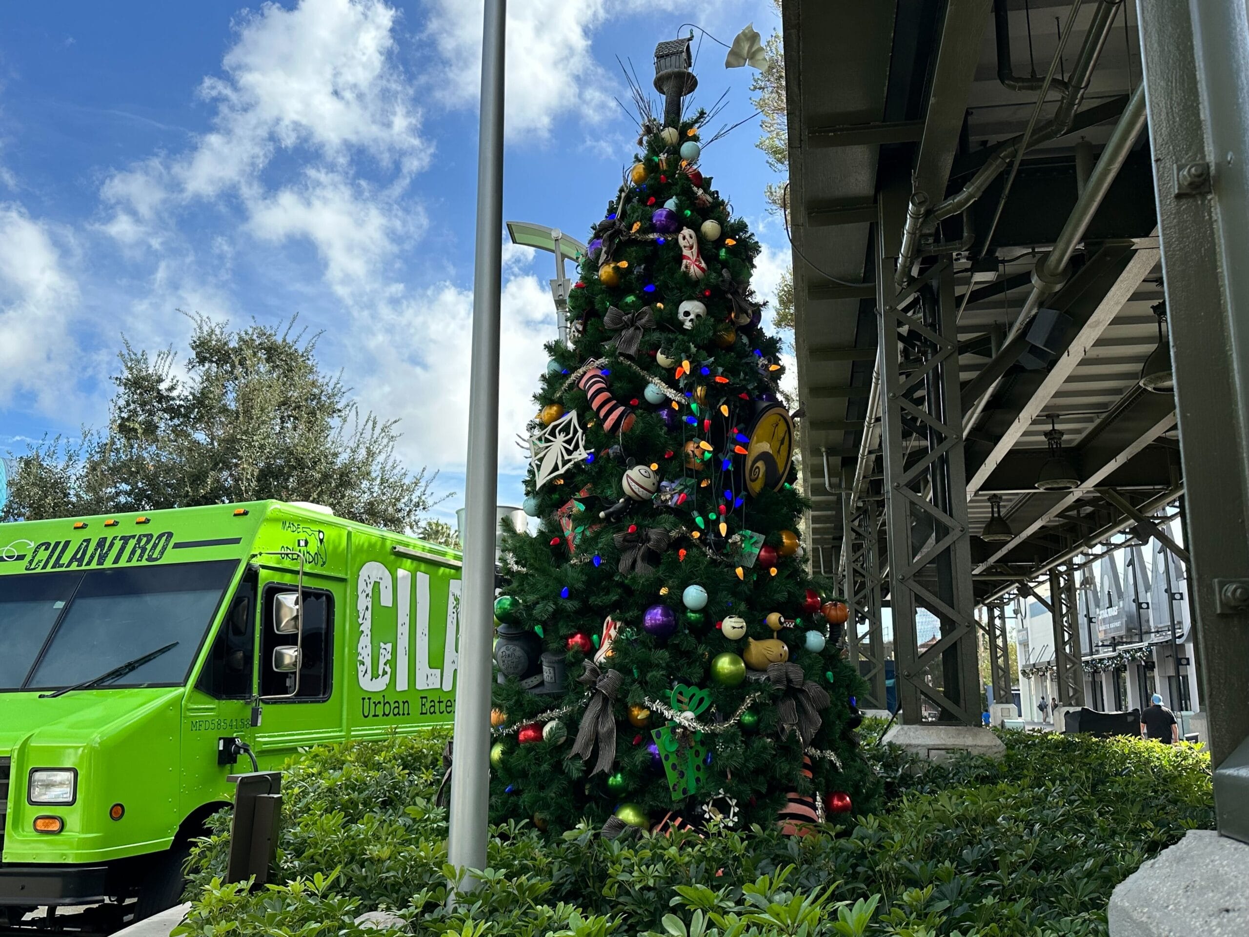 A decorated Christmas tree stands under a metal structure, surrounded by greenery. A bright green food truck is parked nearby.