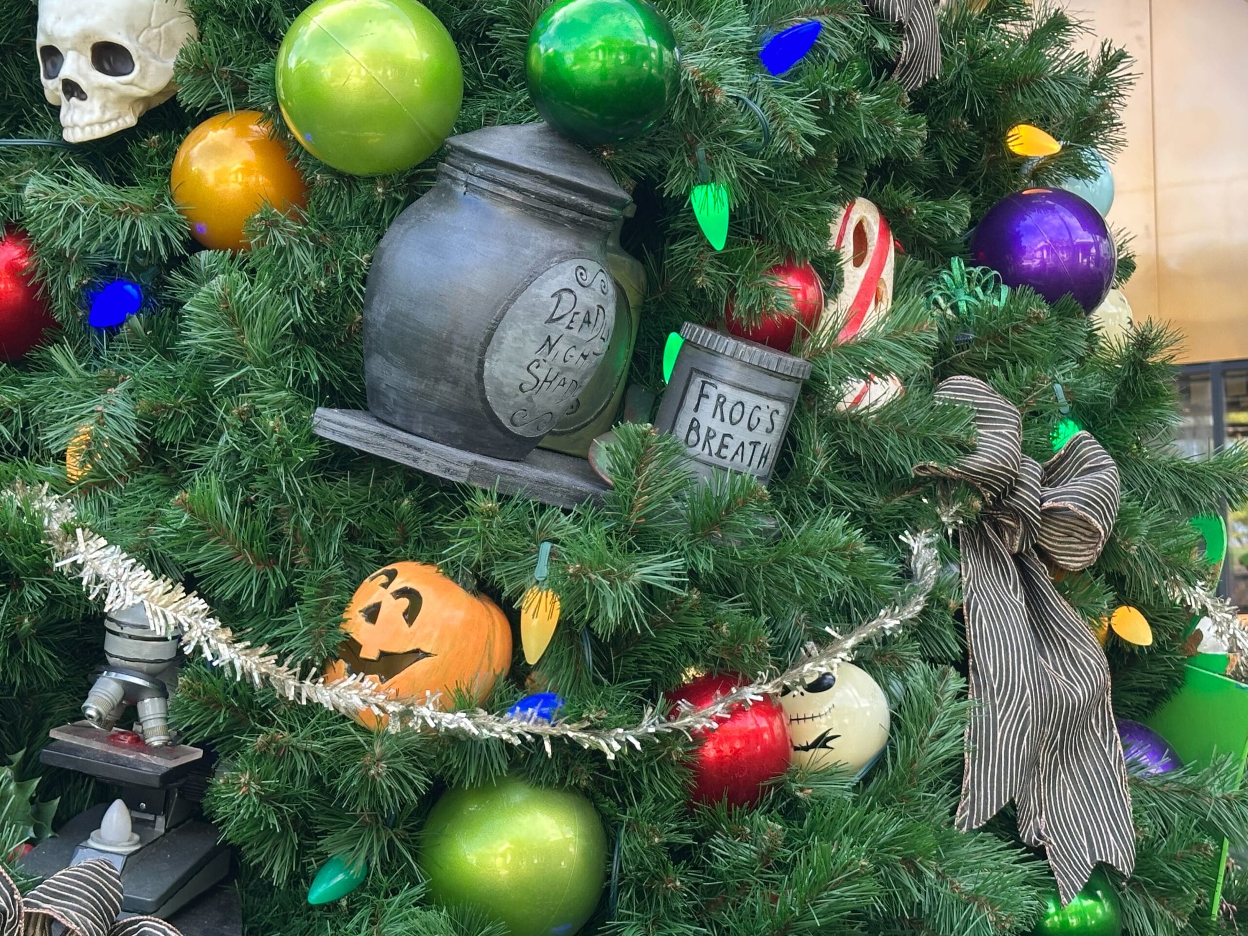 A festive Christmas tree decorated with colorful ornaments, a black urn, a jar labeled "Frog's Breath," and Halloween-themed items like skulls and pumpkins.
