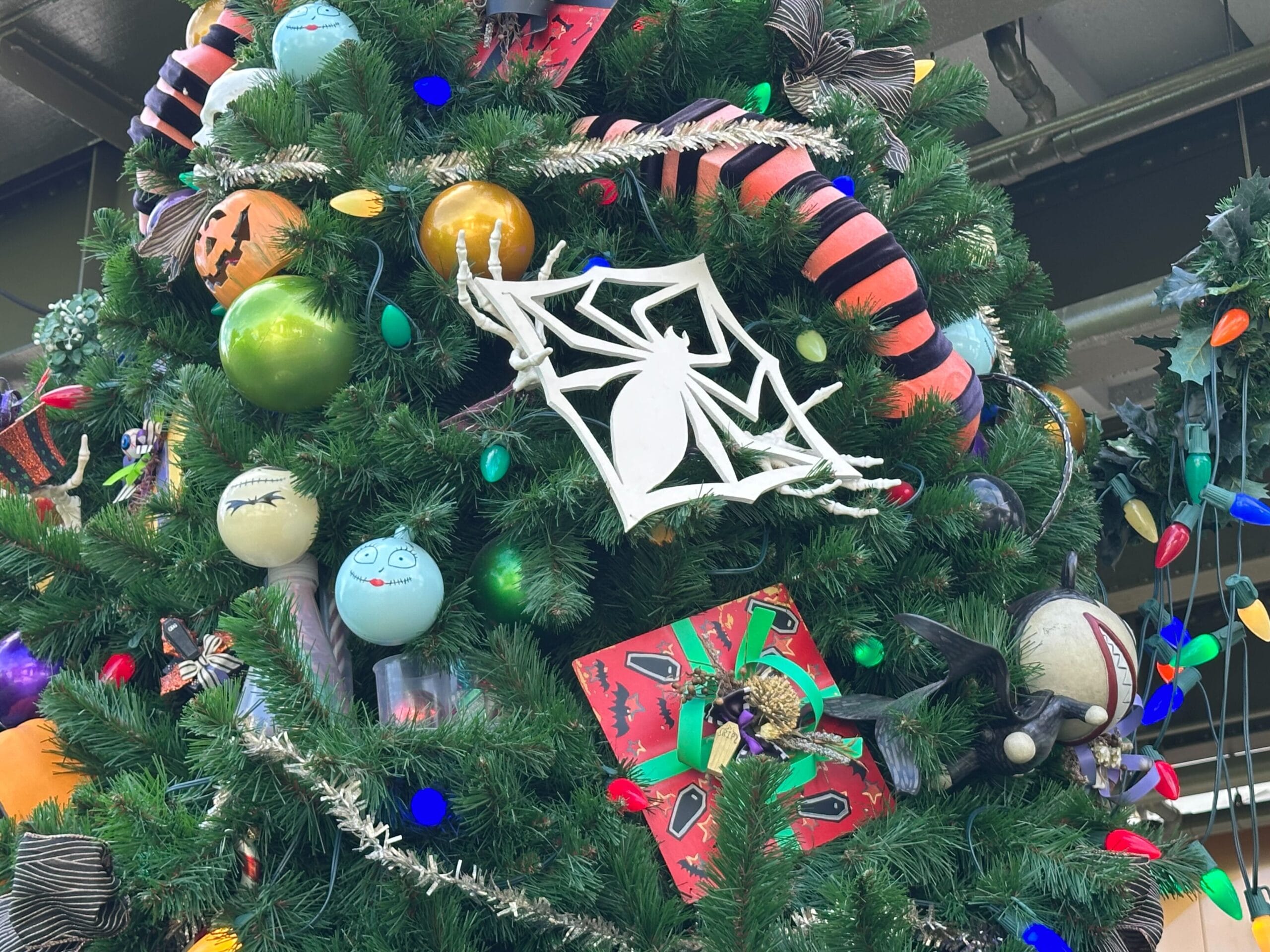 A Christmas tree decorated with Halloween-themed ornaments, including a spider web, colorful balls, and striped ribbon, adorned with multicolored string lights.