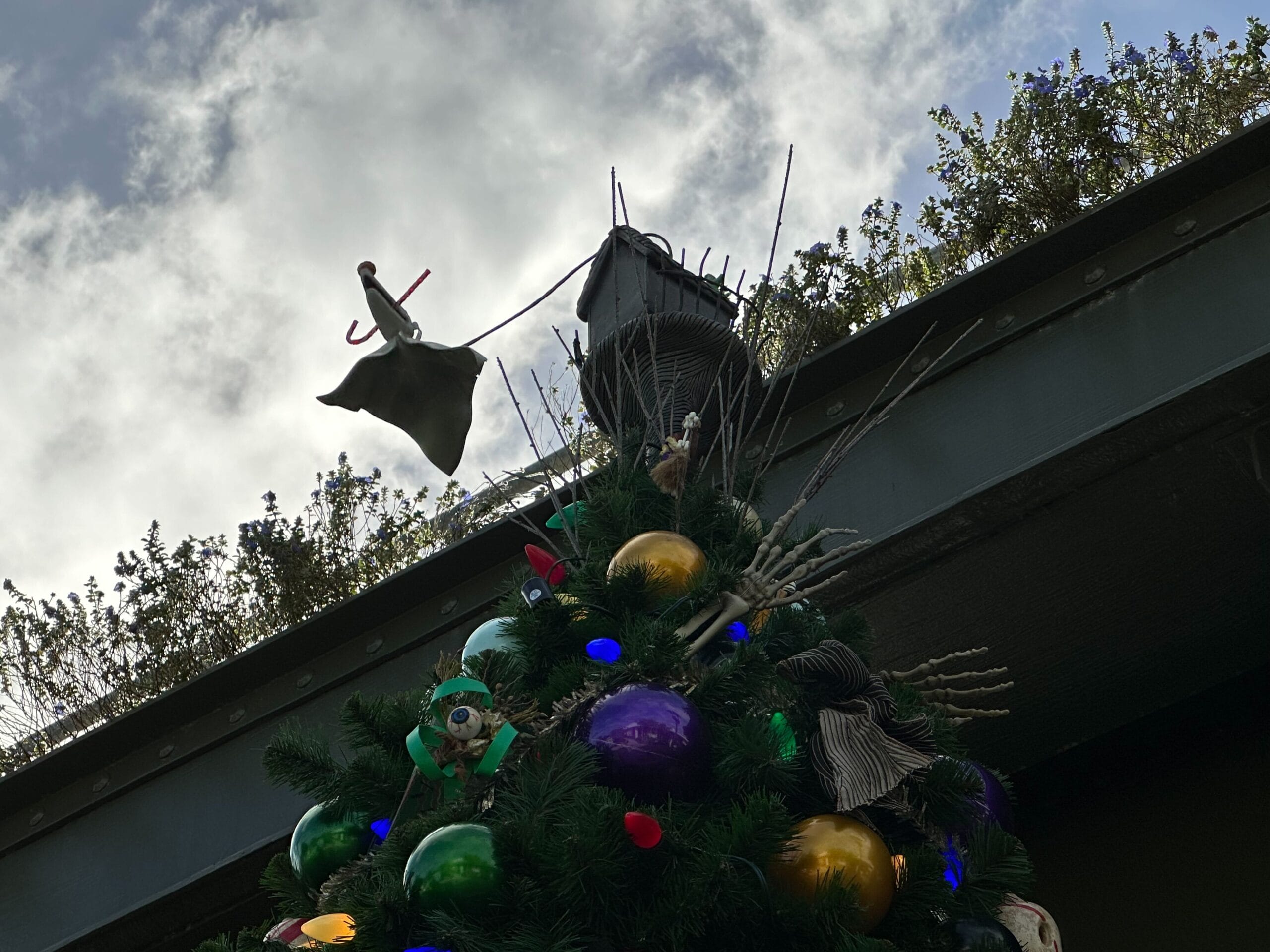 Christmas tree decorated with colorful bulbs and Halloween-themed ornaments, topped with a figure holding a bag, set against a cloudy sky background.