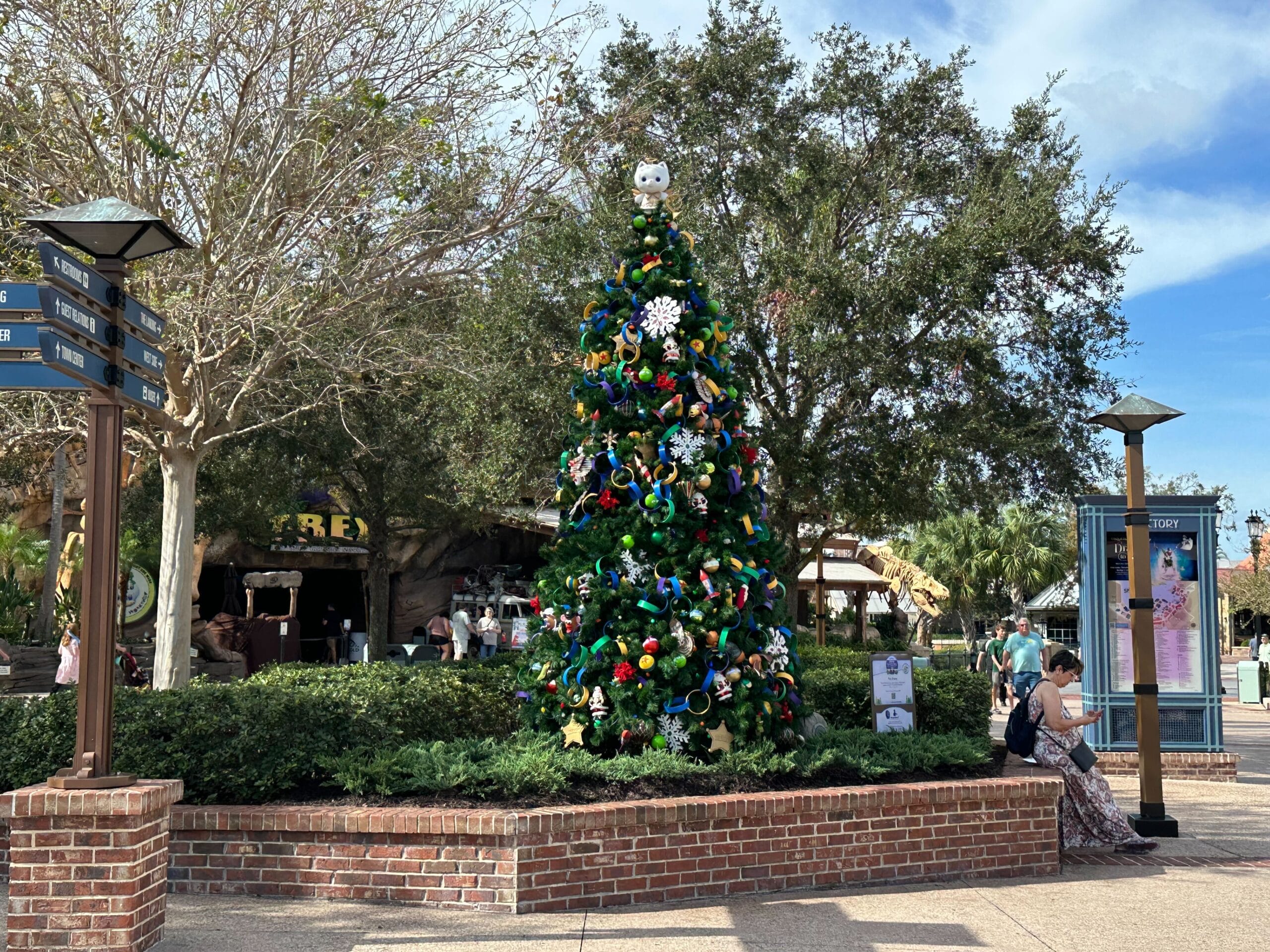 A Christmas tree decorated with colorful ribbons and ornaments is displayed outdoors in a brick planter. People are seated nearby, and trees surround the area under a clear sky.