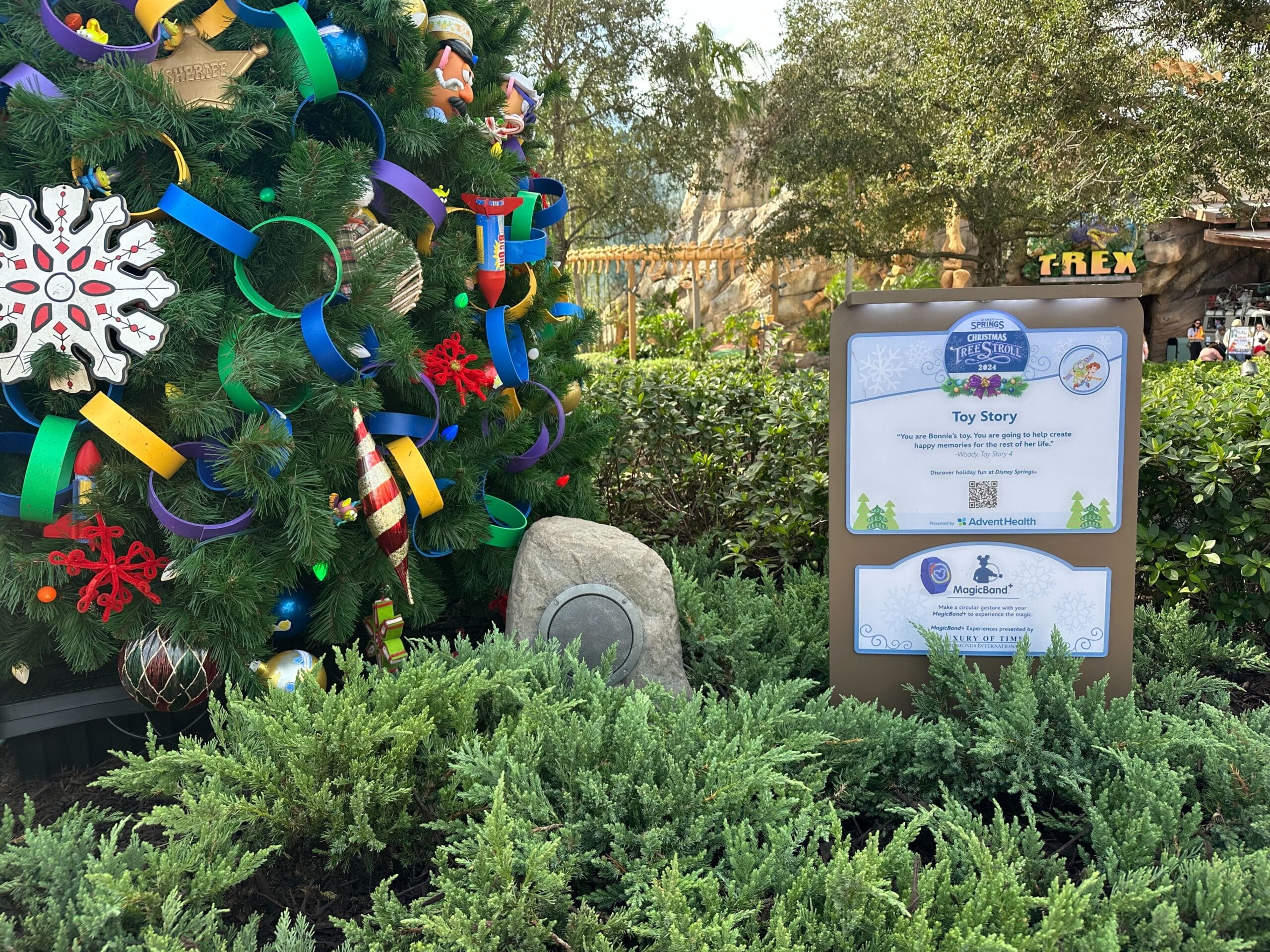 A decorated Christmas tree stands beside a sign titled "Toy Story" in a garden setting with greenery and a brown rock.