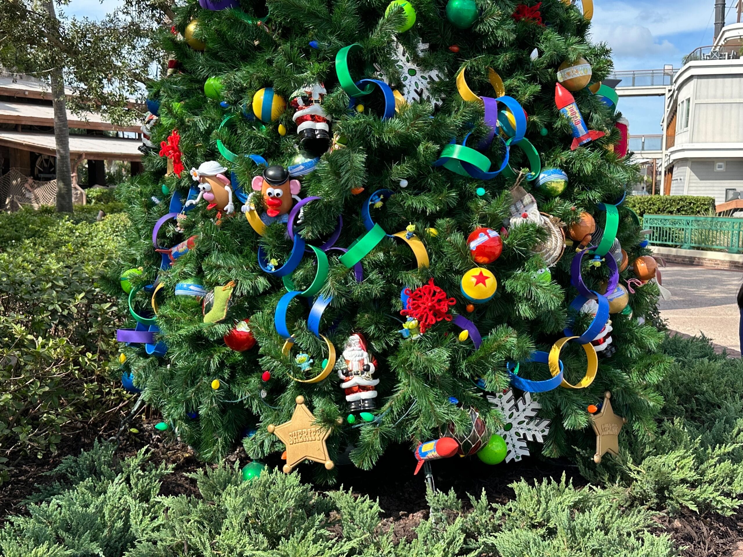 A Christmas tree adorned with colorful ribbons, ornaments, and small toy figures is set outdoors with greenery in the background.