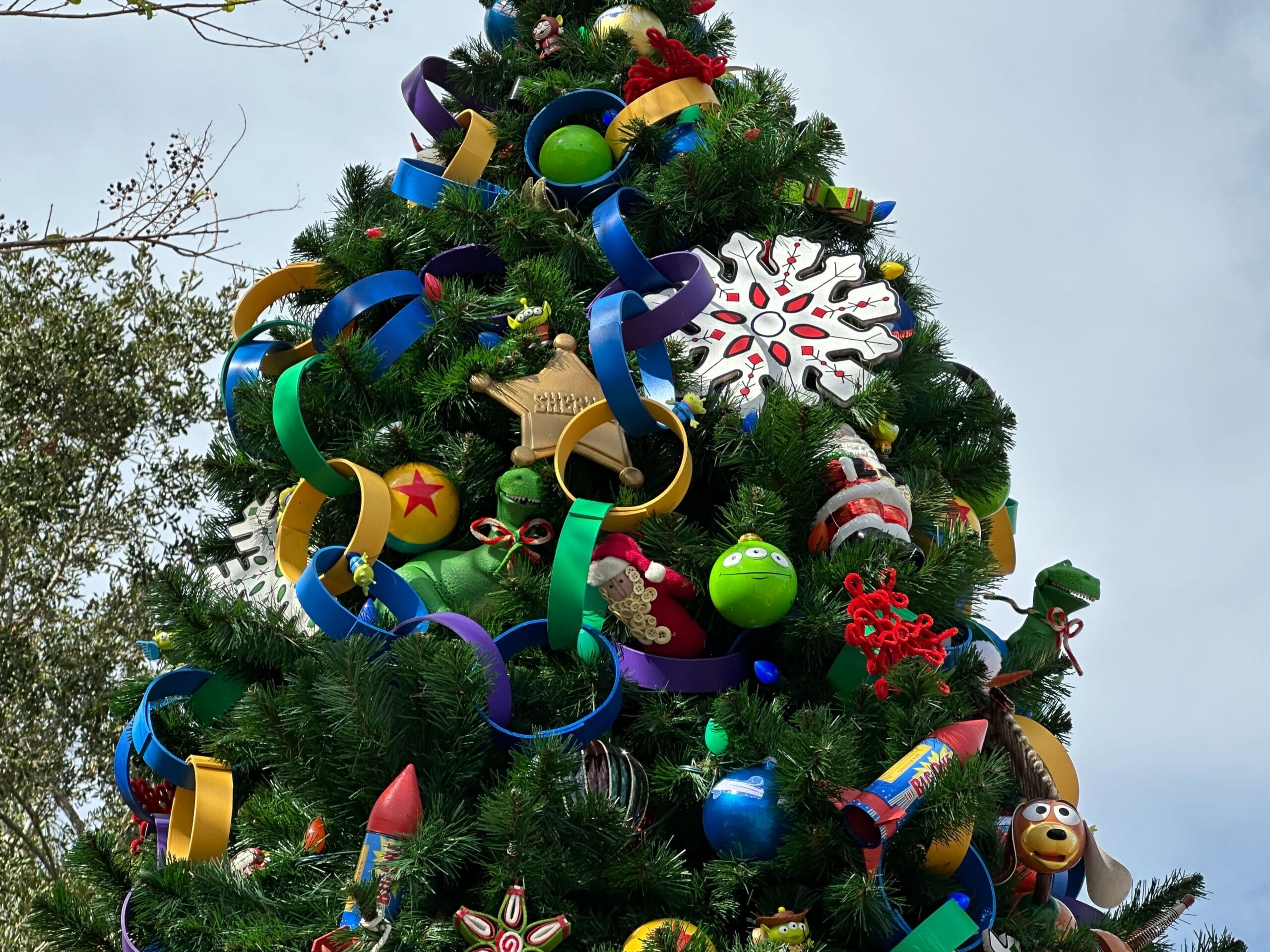 A tall Christmas tree decorated with colorful ornaments, ribbons, and toy-themed decorations against a cloudy sky.