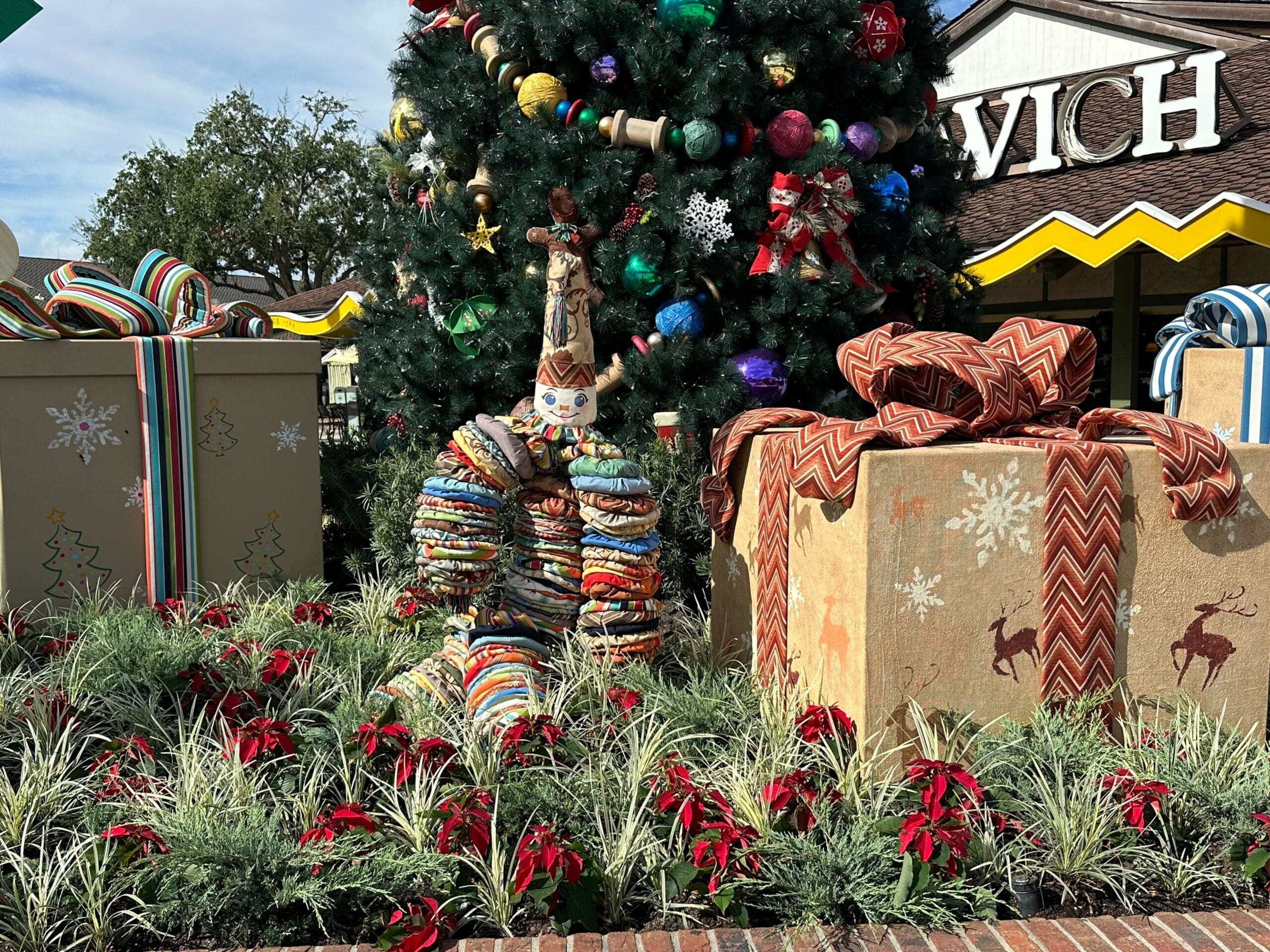 A decorated Christmas tree with large wrapped gifts and a whimsical stack of donuts centerpiece in a landscaped setting.