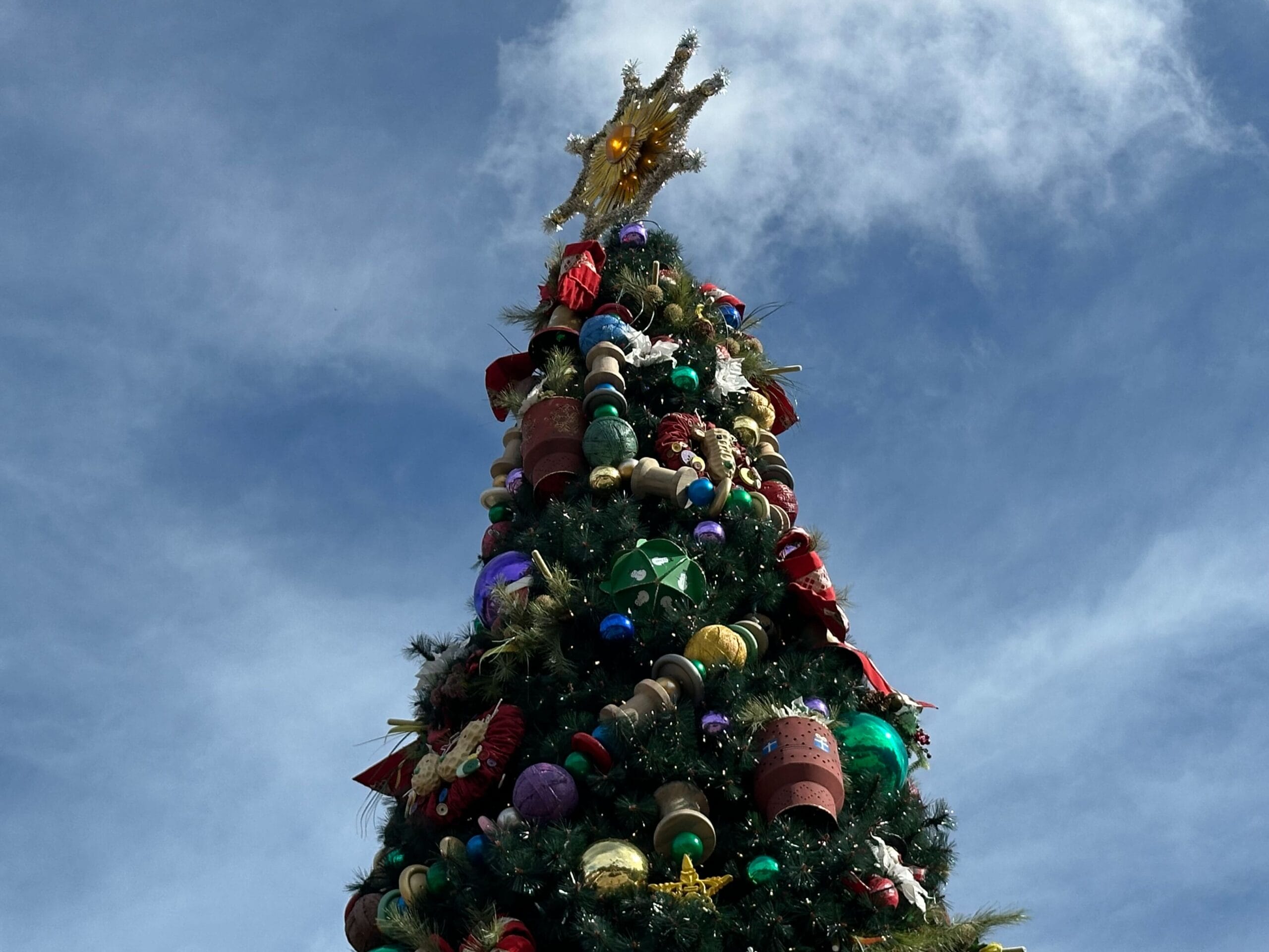 Tall Christmas tree adorned with colorful decorations and a star on top, set against a blue sky with clouds.