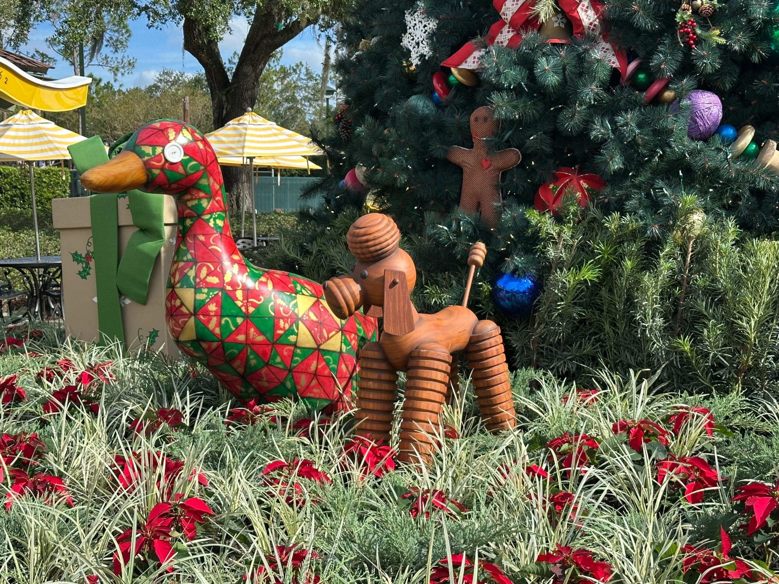 Decorative duck, dog, and gingerbread figure in front of a Christmas tree with ornaments, surrounded by greenery and red poinsettias.