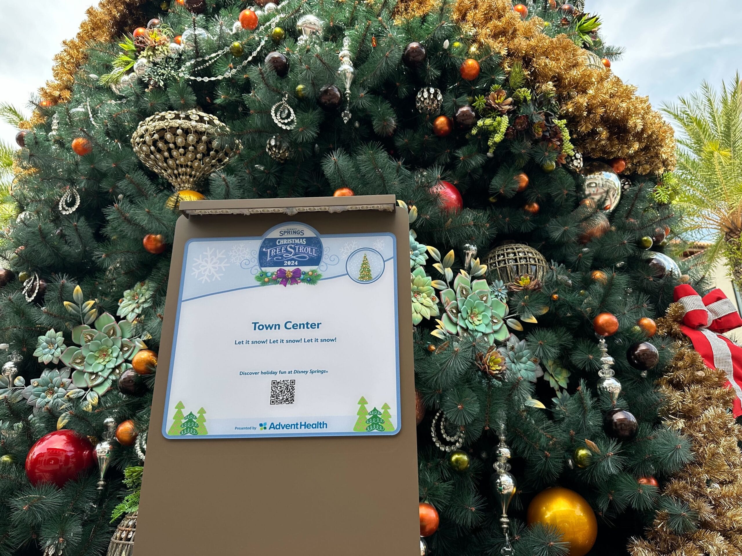 In front of a grand, decorated Christmas tree stands a sign that reads "Christmas Tree Stroll - Town Center," highlighting the magic of Disney Springs and proudly sponsored by AdventHealth.