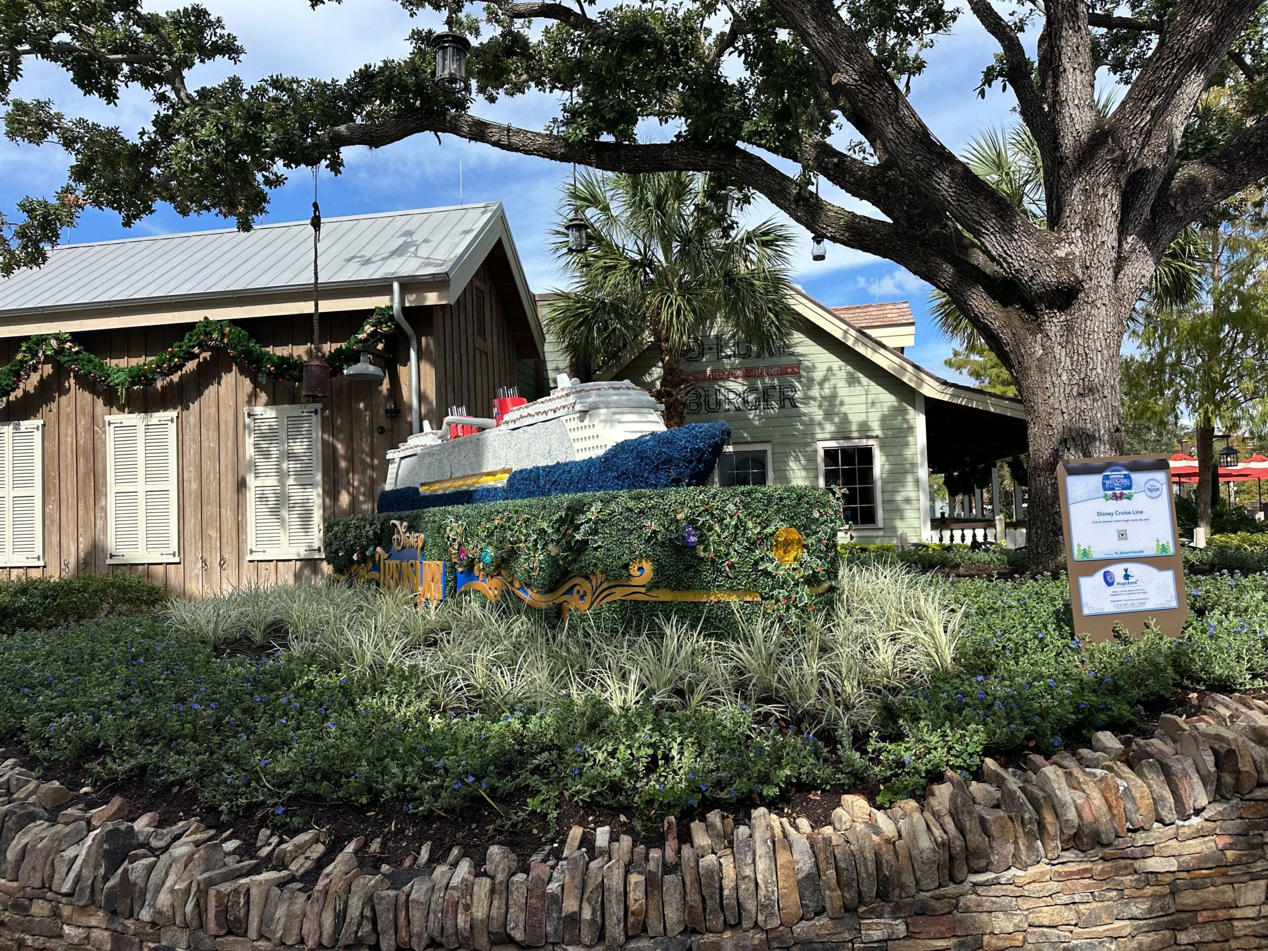 Model of a cruise ship displayed outdoors, surrounded by greenery and a stone wall, near a wooden building with holiday decorations.