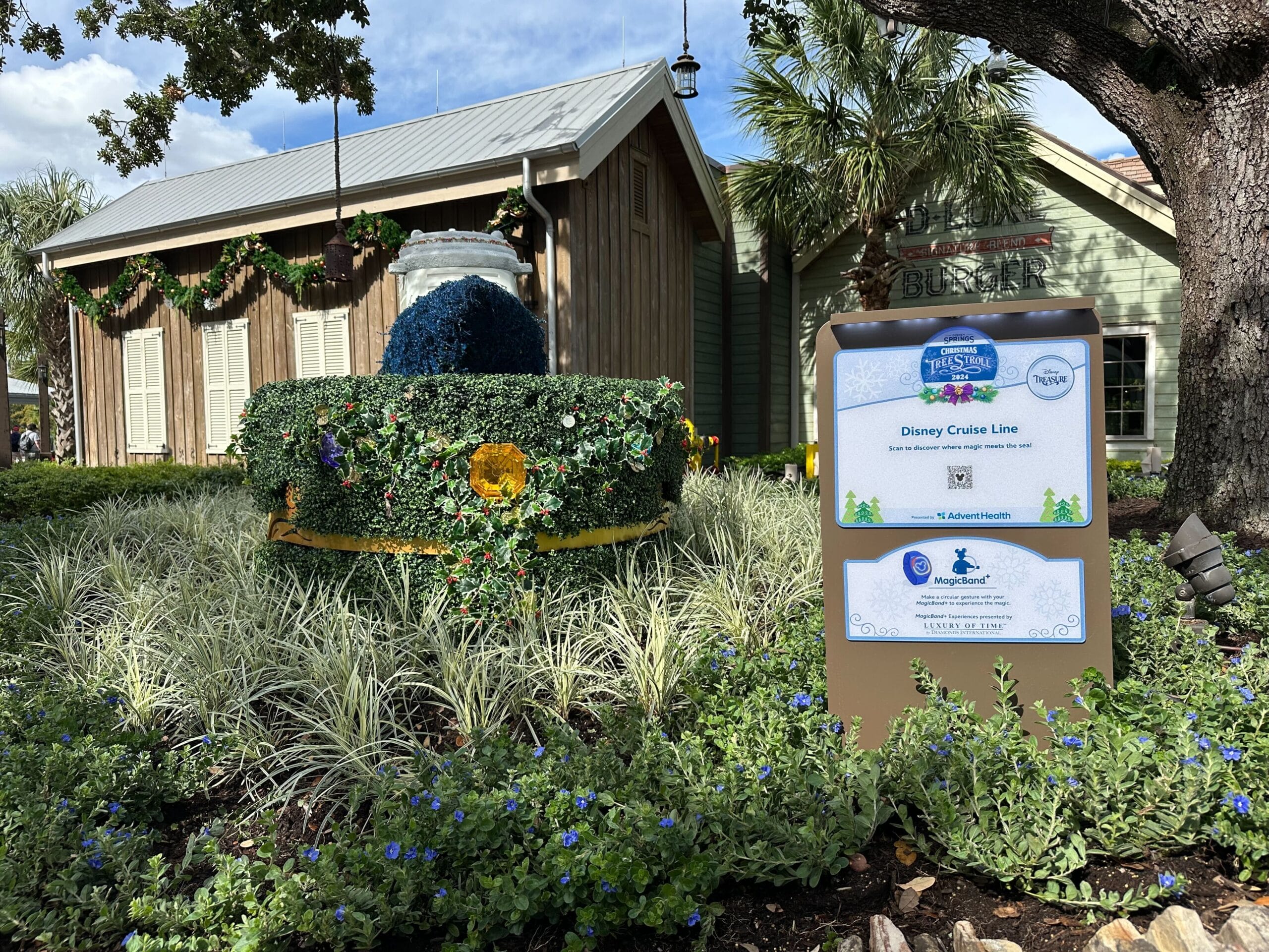 A Disney-themed flower garden with a decorated hat sculpture is situated near a building. A sign about the Disney Cruise Line is in the foreground.