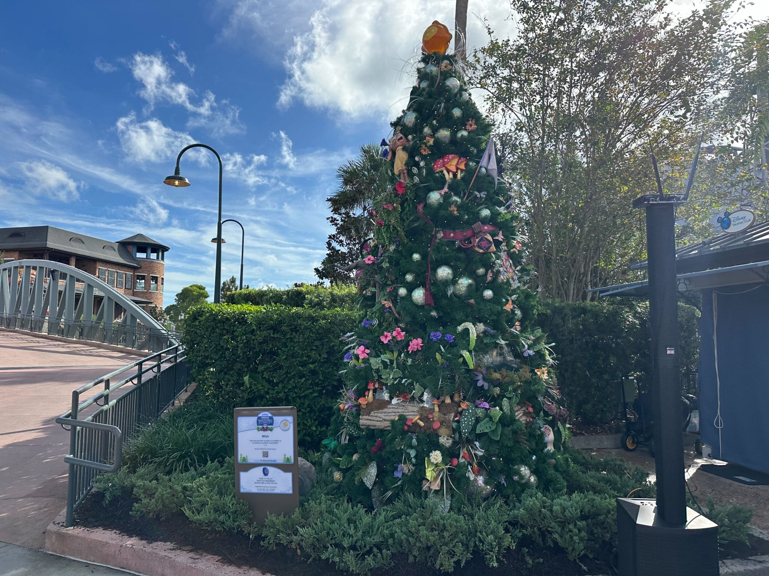 Decorated Christmas tree outdoors near a lamp post and bridge, with greenery and a sign nearby.