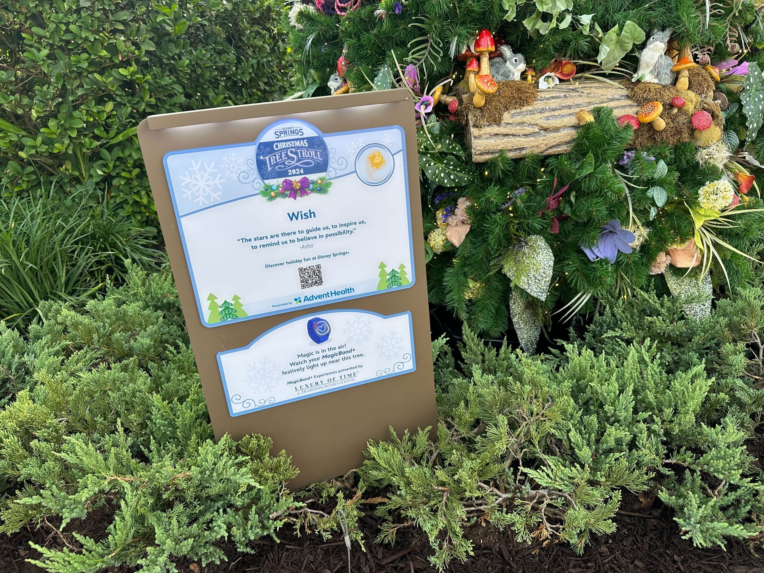 Sign next to a decorated Christmas tree displays a QR code and a message titled "Wish," set among greenery in an outdoor setting.