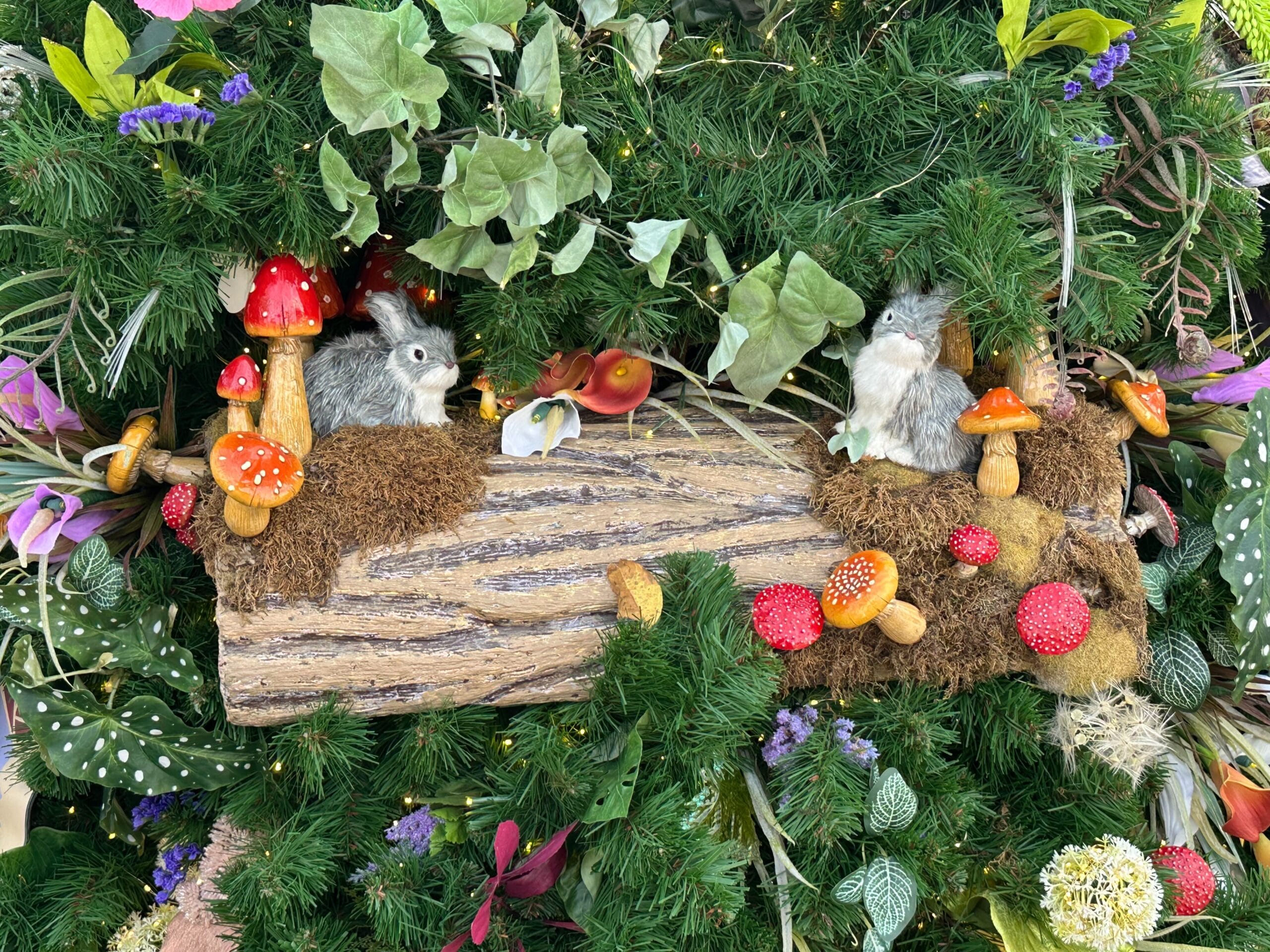 Decorative display featuring two plush rabbits on a log surrounded by assorted mushrooms, greenery, and purple flowers.