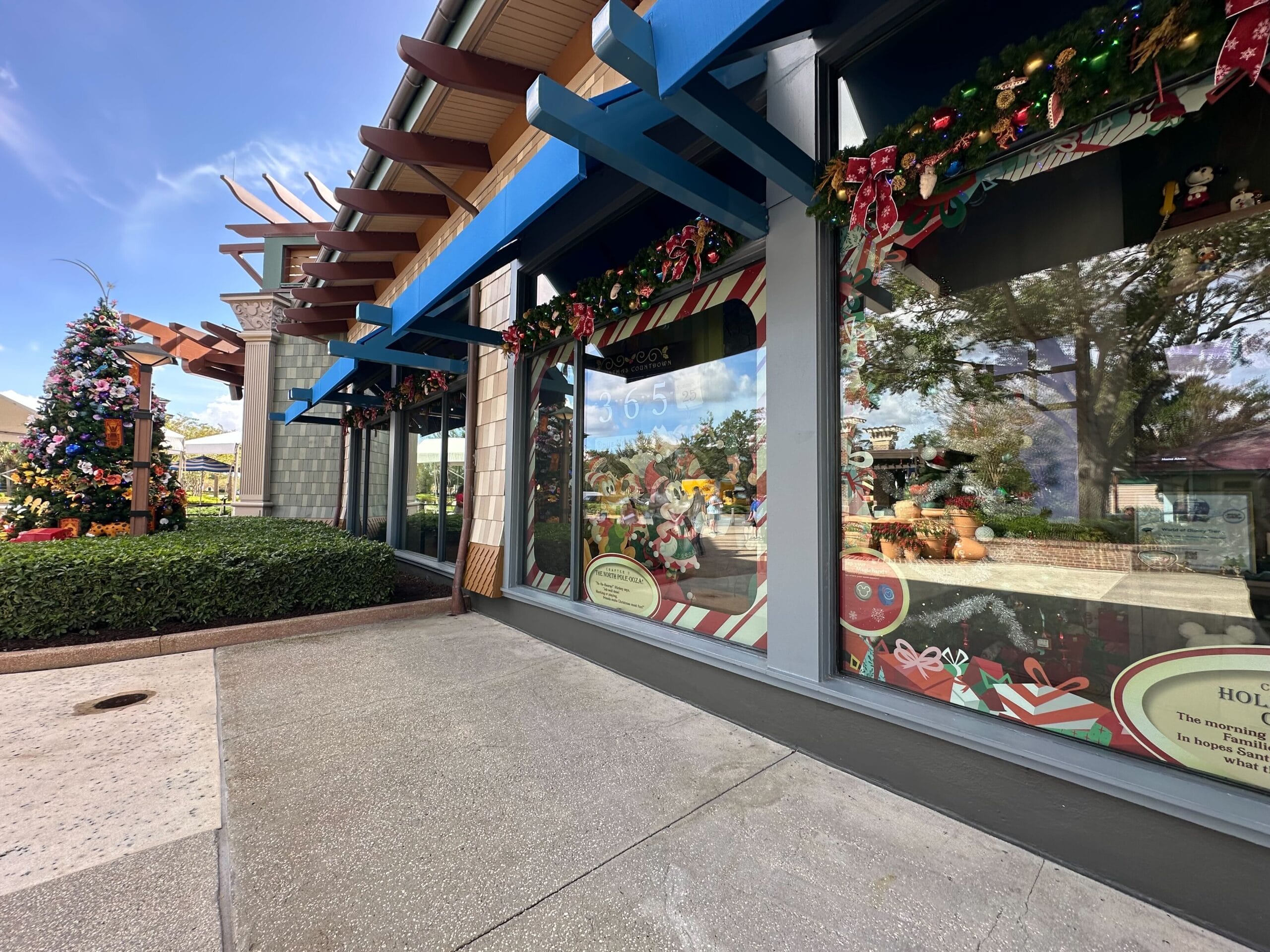 Storefront decorated with festive holiday ornaments and garlands. A colorful tree stands outside, with a path leading past greenery under a blue sky.