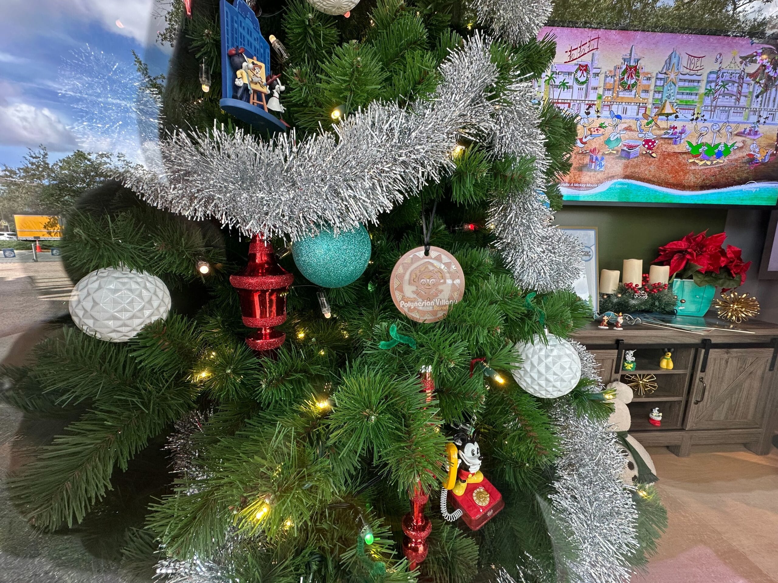 Close-up of a decorated Christmas tree with tinsel, white and red ornaments, and a character ornament. A television and candles are in the background.