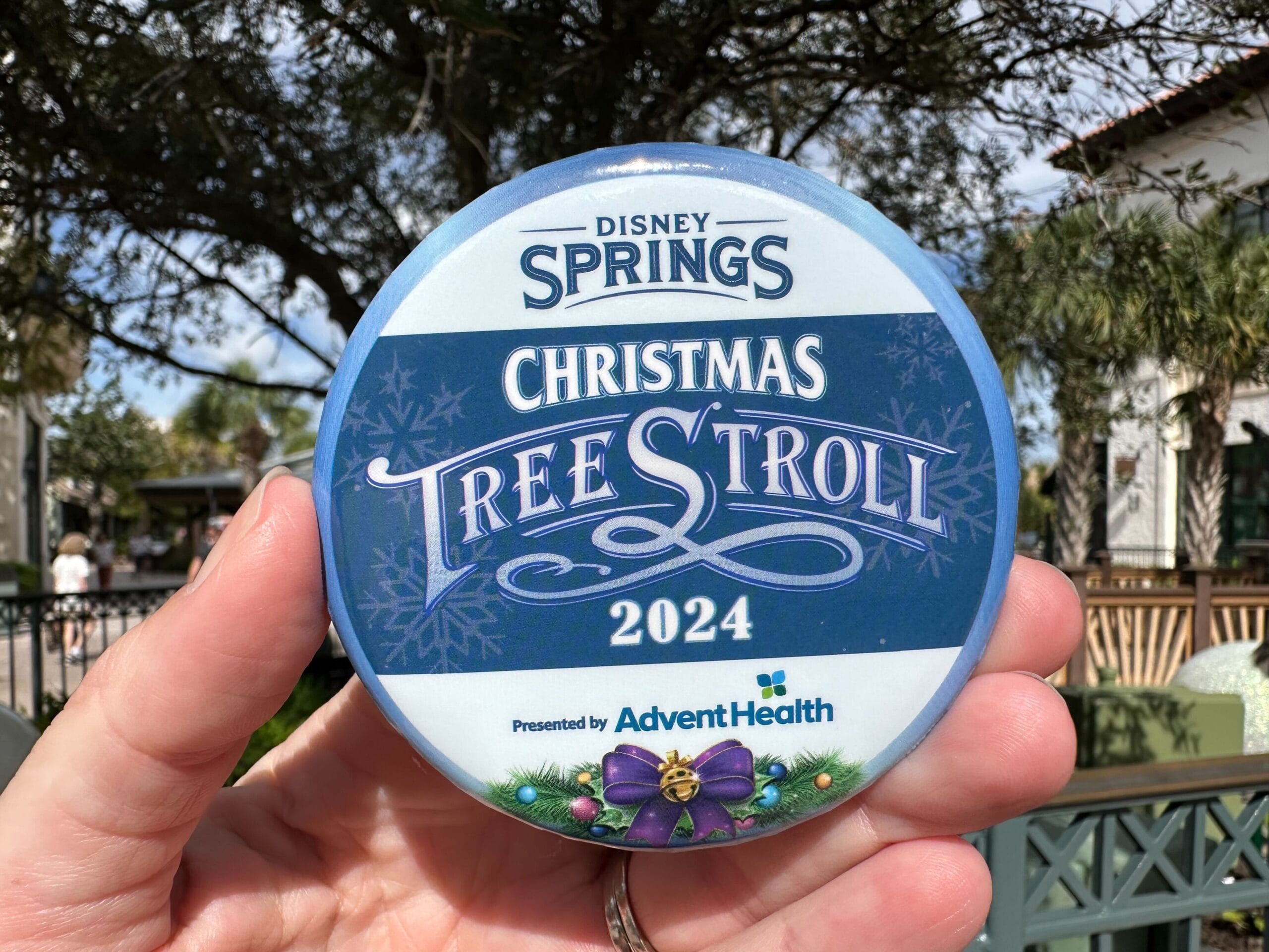 A hand holds a button featuring the "Disney Springs Christmas Tree Stroll 2024" text alongside the Advent Health logo, glistening under the warm sunshine outdoors.
