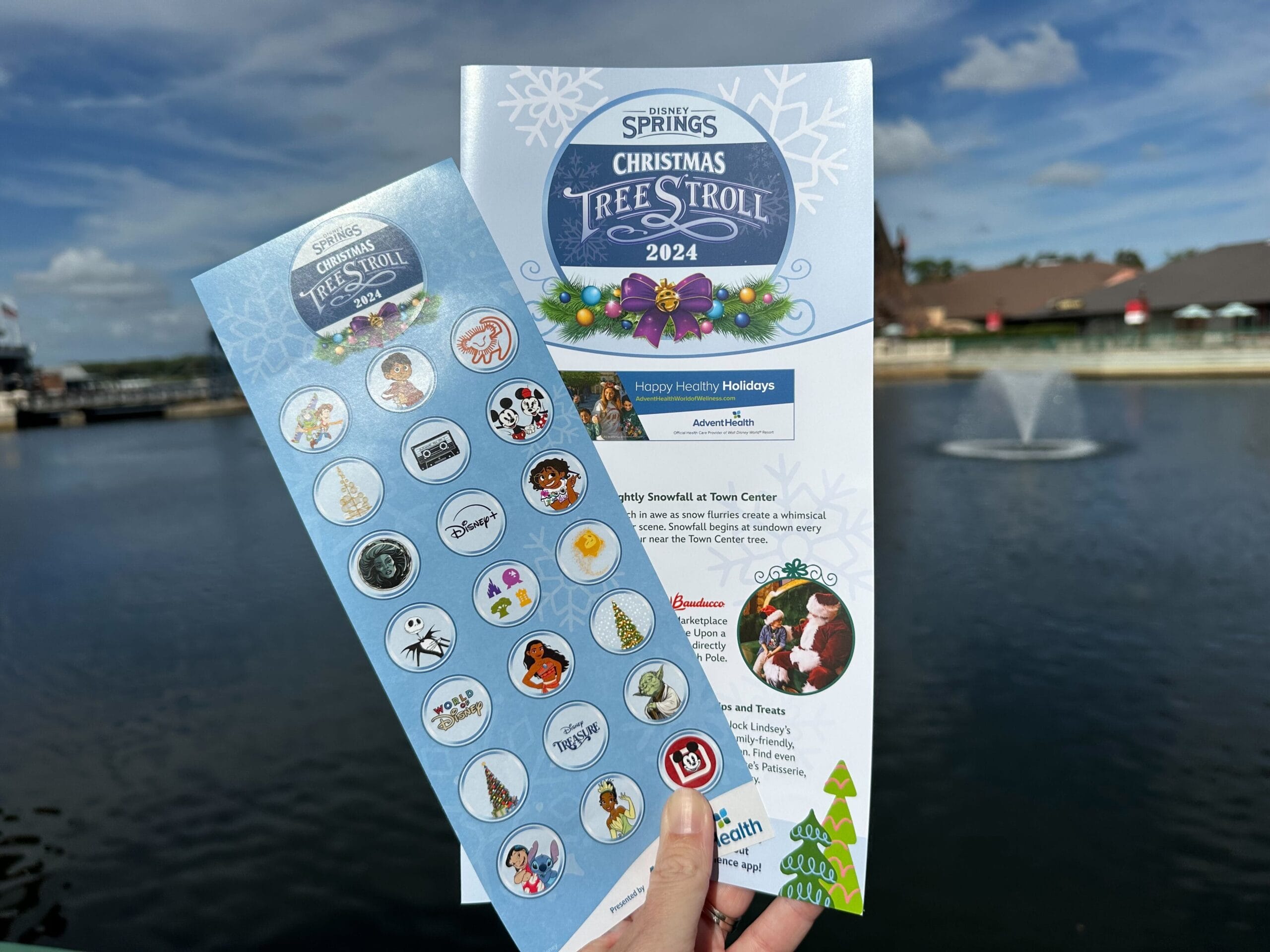 A hand holds the 2024 Disney Springs Christmas Tree Stroll map and a festive sticker sheet beside a shimmering lake, complete with a fountain under a bright blue sky.