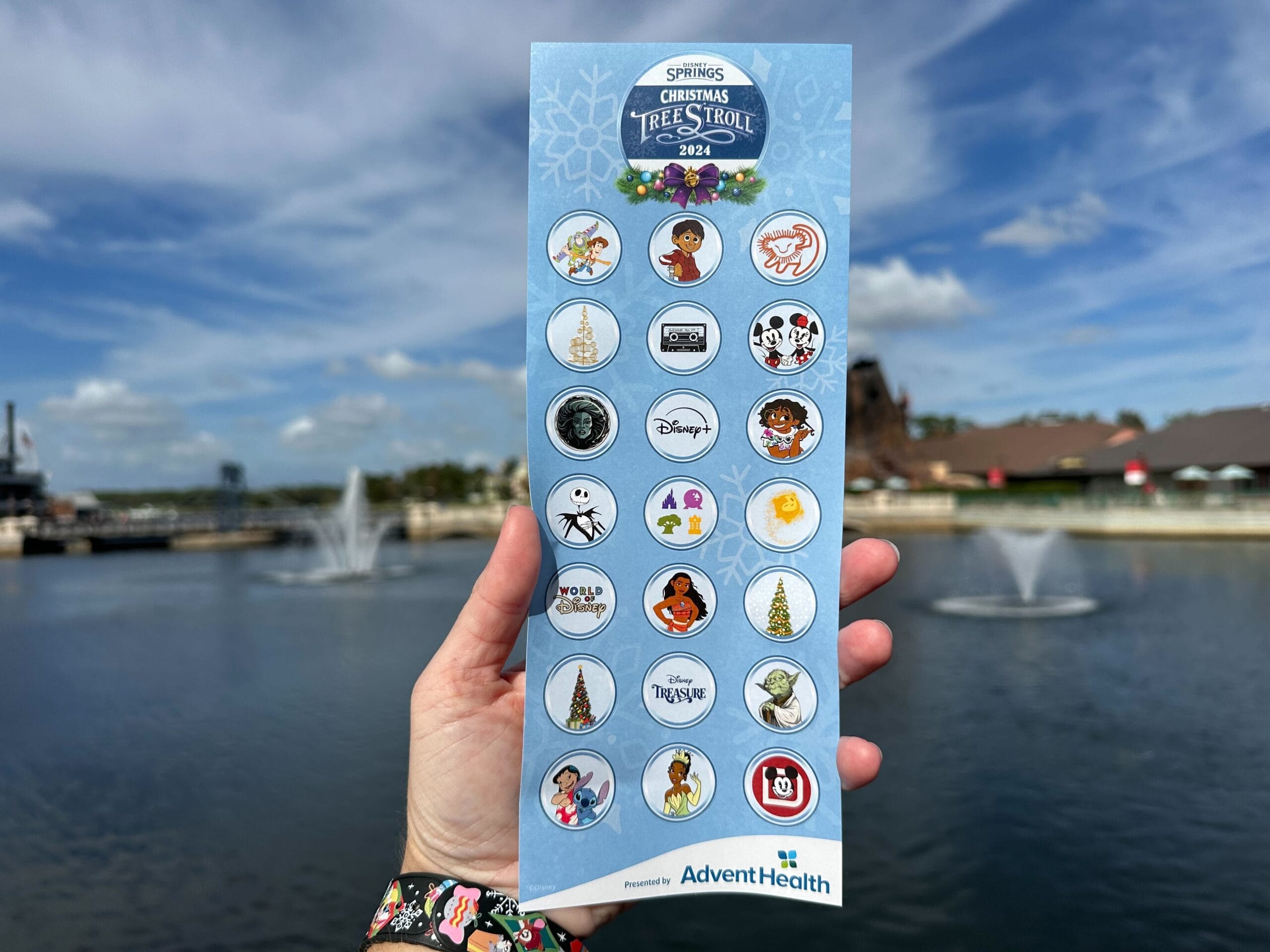 A person clutching their Disney Springs Christmas Tree Stroll map, adorned with themed stickers, stands against a backdrop of fountains under a cloudy sky.