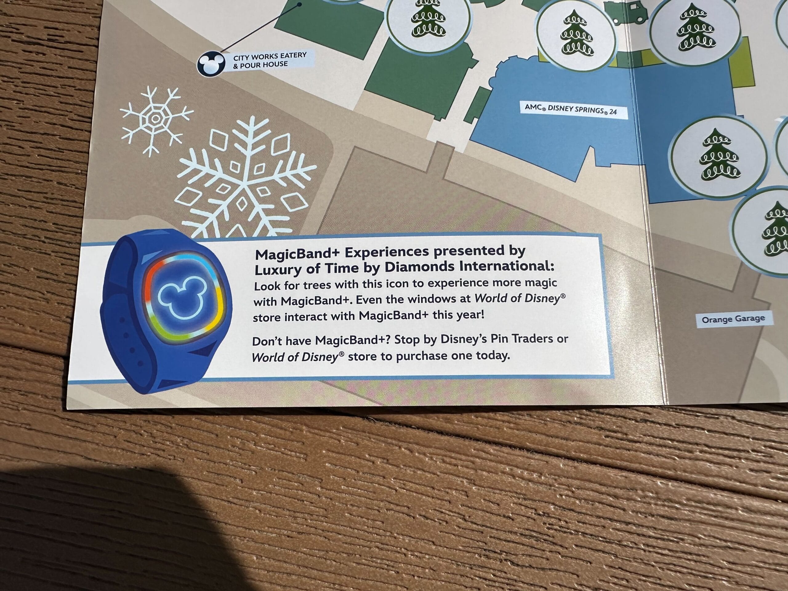 Close-up of a brochure featuring a MagicBand+ ad, showcasing a blue band with a Mickey Mouse logo, nestled on a map background. Icons like snowflakes hint at festive Disney experiences, including the enchanting Disney Springs Christmas Tree Stroll.