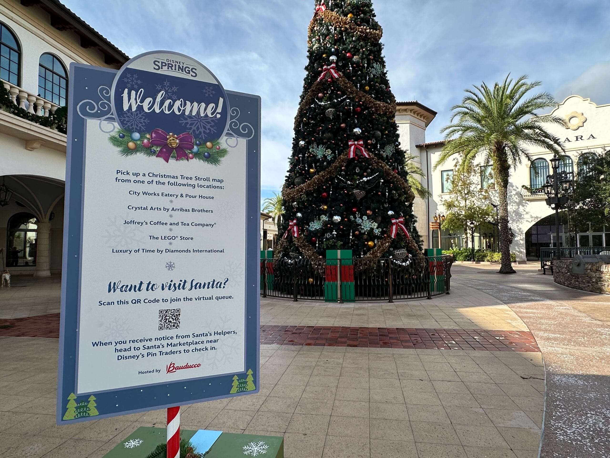 A signboard with instructions stands in front of a large decorated Christmas tree in the plaza, reminiscent of the Disney Springs Christmas Tree Stroll. Nearby buildings feature arched windows, and palm trees grace the background, creating a festive blend of tradition and tropical charm.