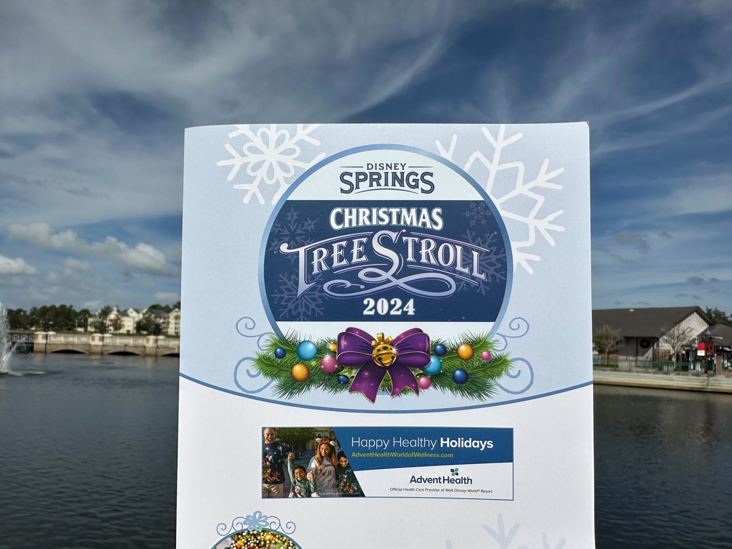 Amid holiday decorations and a snowy lakeside backdrop, a person holds a festive "Disney Springs Christmas Tree Stroll 2024" flyer, inviting all to embark on this enchanting seasonal adventure.