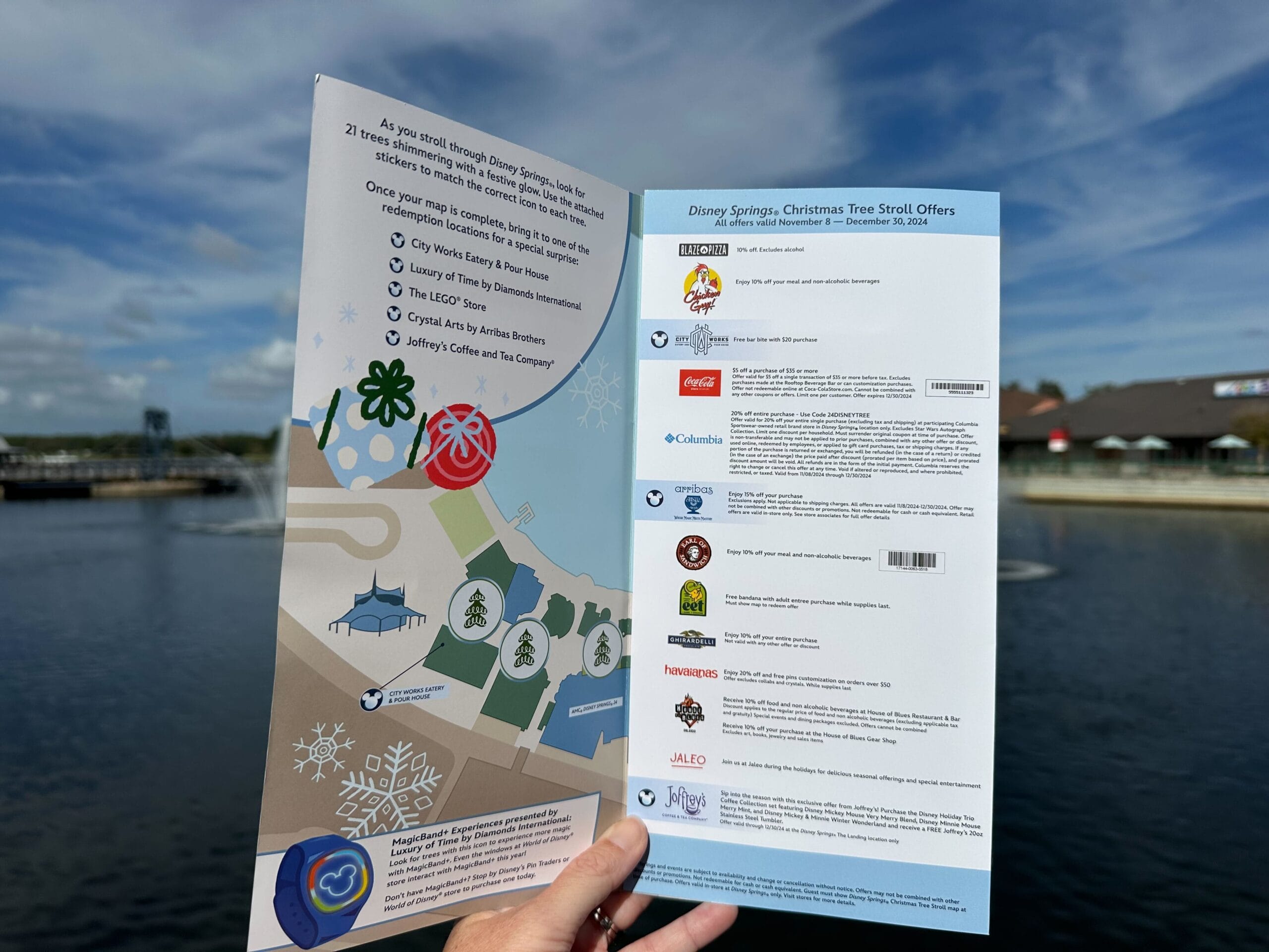 A hand holds an open brochure against a waterfront background, showcasing the Disney Springs Christmas Tree Stroll. On the left is a festive map guiding you through enchanting trees, while the right page highlights special offers.