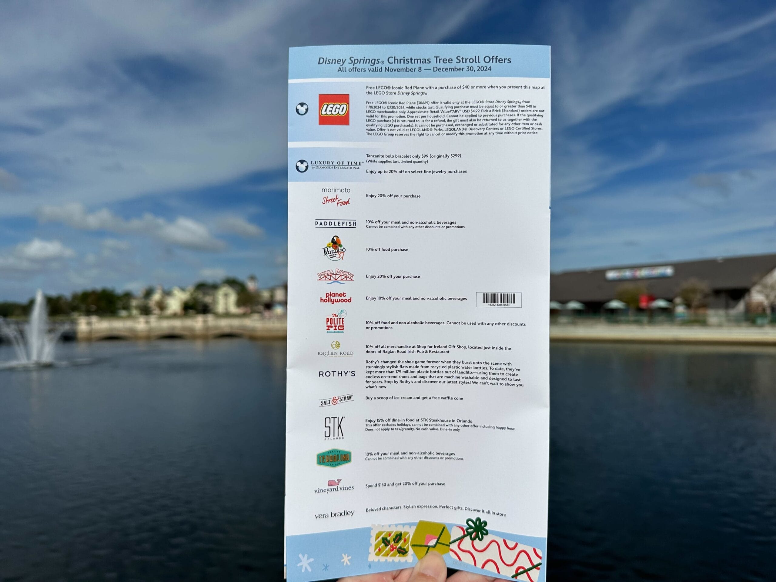 A hand holds a brochure titled "Disney Springs: Christmas Tree Stroll Offers" in front of a lake, where twinkling shops and gentle fountains create a festive backdrop. Various brand logos and seasonal offers are prominently listed on the vibrant brochure.