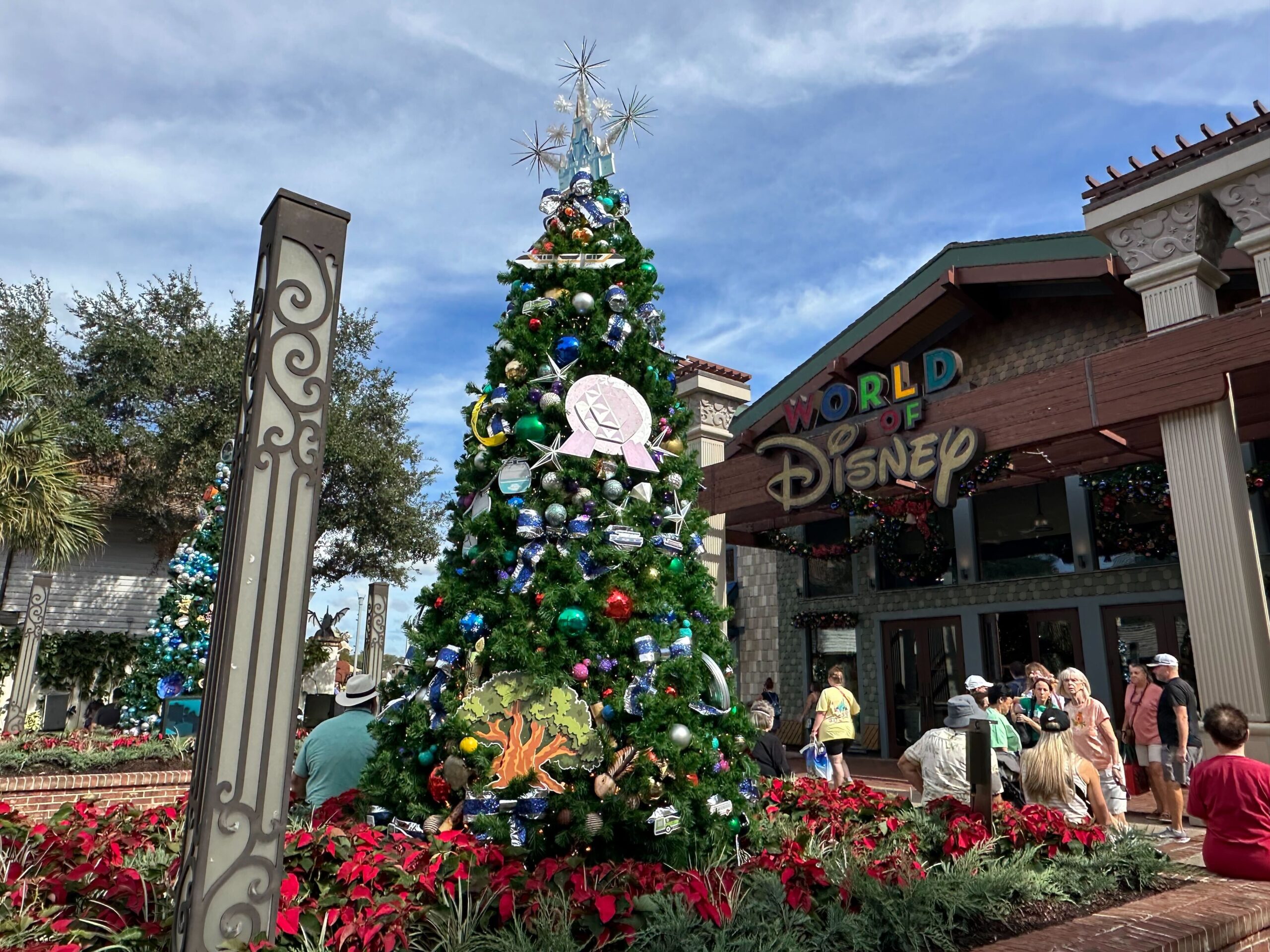 Christmas tree decorated with Disney-themed ornaments in front of the "World of Disney" store. People are walking around, and the sky is partly cloudy.