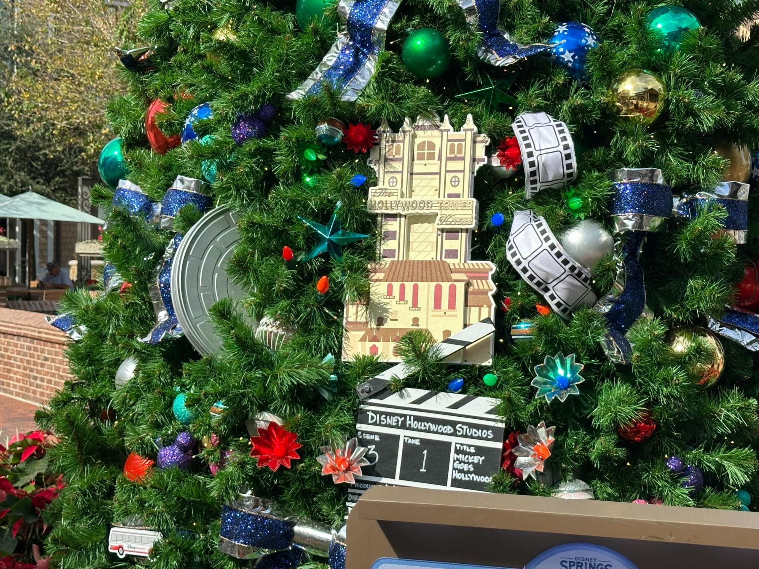 Close-up of a Christmas tree decorated with film-themed ornaments, including a model of a building, a film reel, and a clapboard labeled "Disney Hollywood Studios.