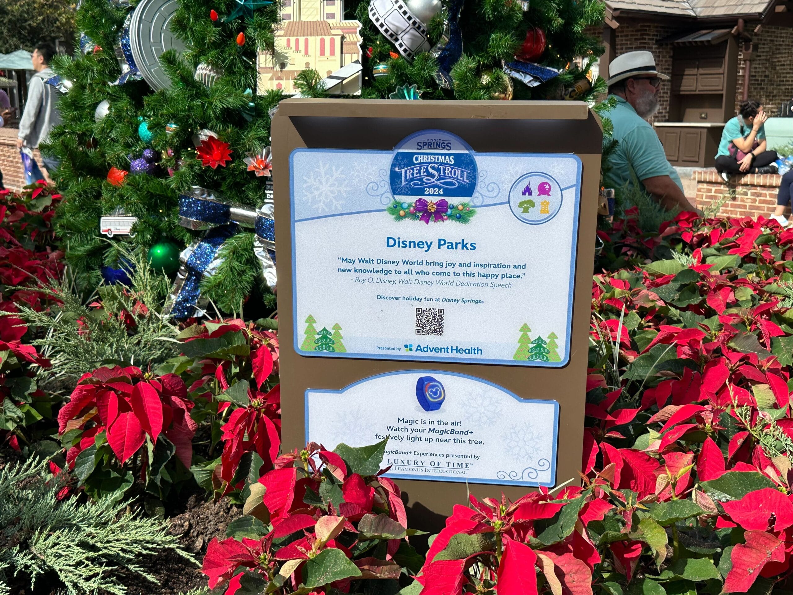 Sign at Disney Parks' Christmas Tree Stroll 2022, surrounded by poinsettias and festive decorations, with a QR code and quote.