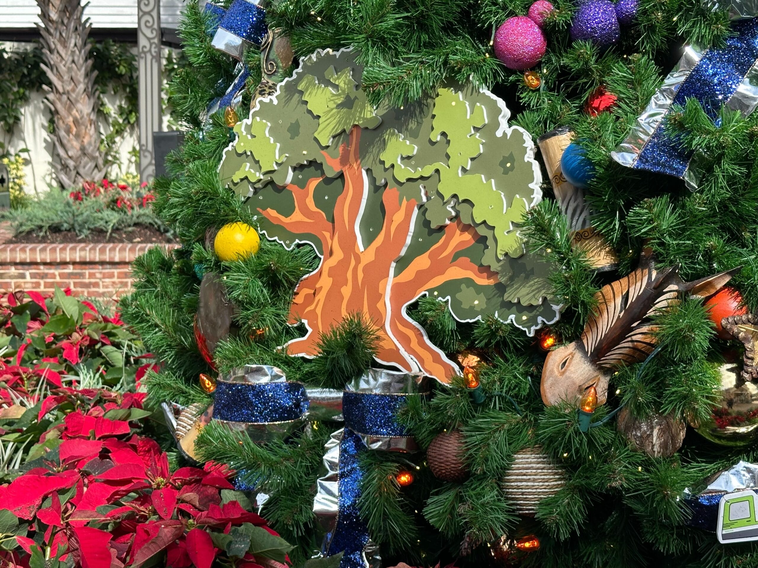 Christmas tree with a painted wooden tree ornament, surrounded by blue ribbons, colorful baubles, and poinsettias at the base.