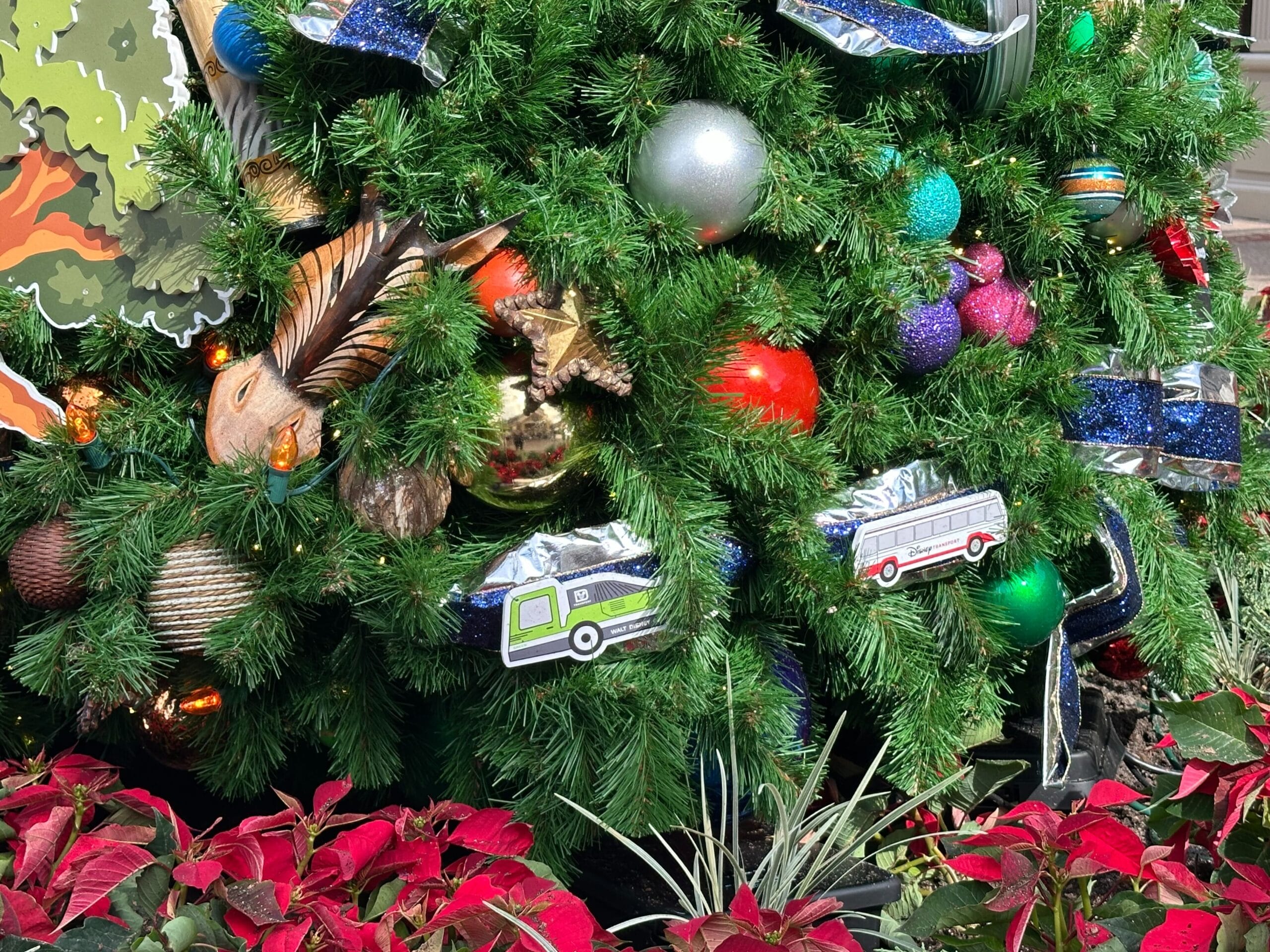 Close-up of a Christmas tree decorated with shiny ornaments and small bus replicas, surrounded by red poinsettias.