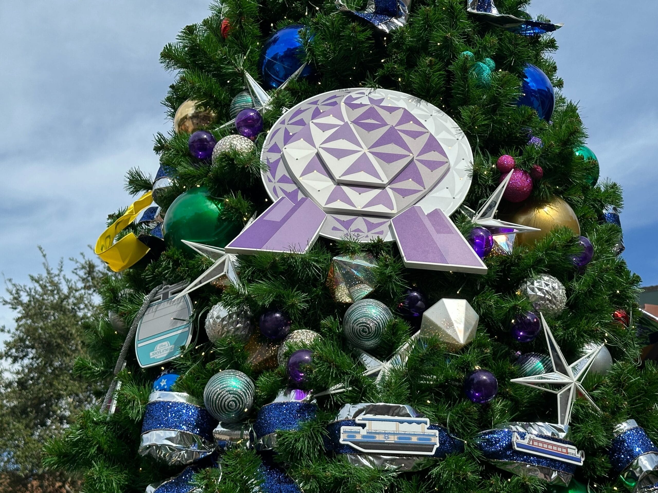 Close-up of a decorated Christmas tree featuring metallic ornaments, geometric shapes, and colorful baubles, with a large purple emblem near the top.