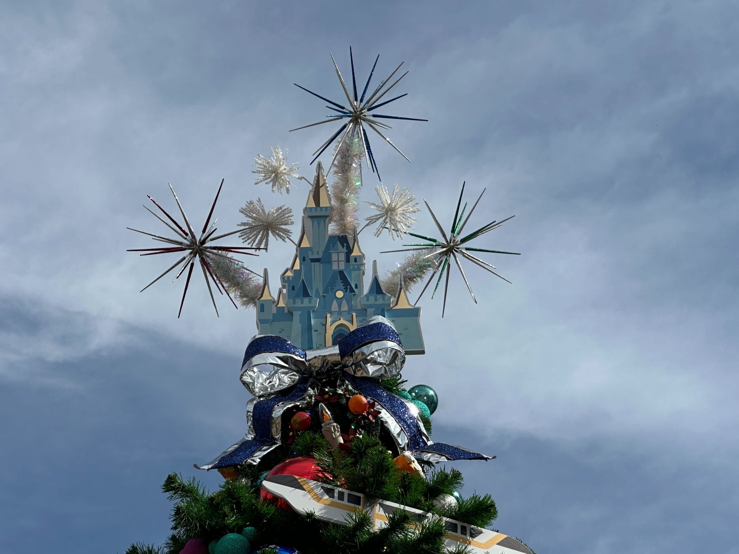 A decorated Christmas tree with a model castle and star-shaped ornaments on top against a cloudy sky.