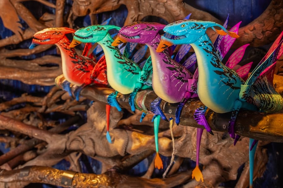 Four colorful dragon-like creatures with wings are perched on a branch. Each creature is distinctively colored in red, green, turquoise, and blue.