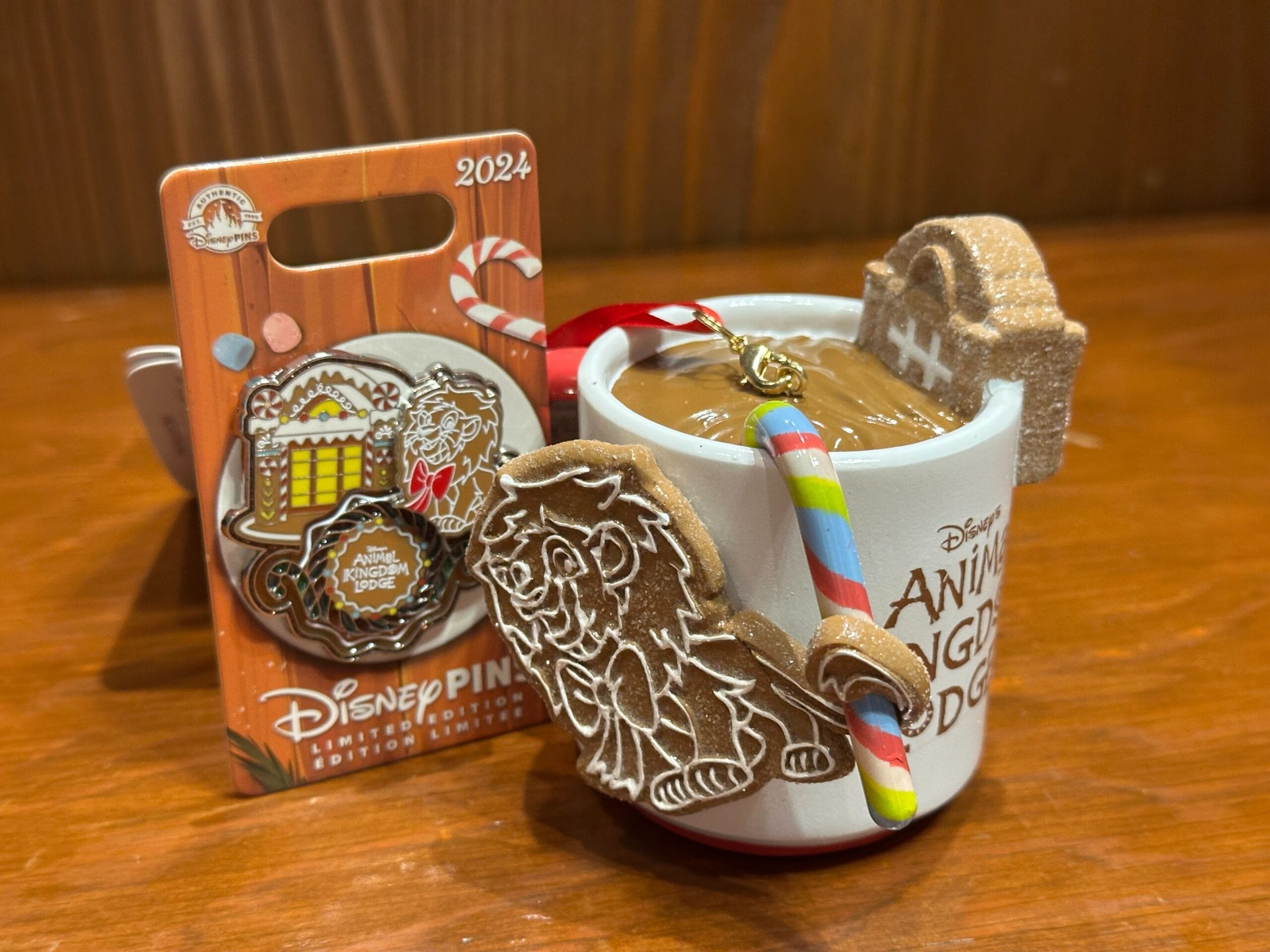 A Disney-themed mug filled with a brown beverage, adorned with a candy cane, lion-shaped cookie, and a gravestone-shaped cookie. A 2024 collectible Disney pin is displayed beside it.