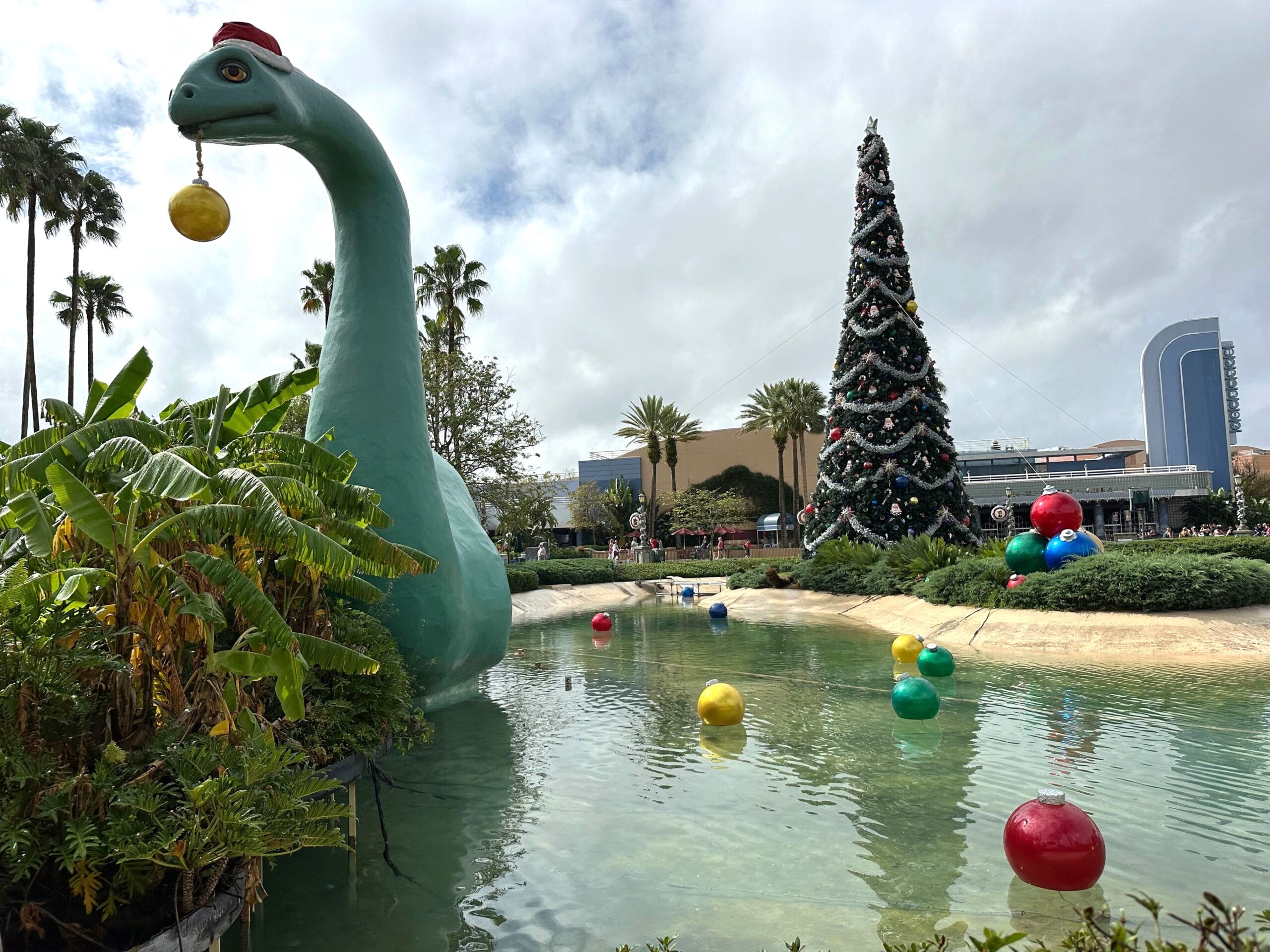 A dinosaur statue holds a bauble in its mouth next to a decorated Christmas tree by a pond with floating colorful spheres under a cloudy sky.