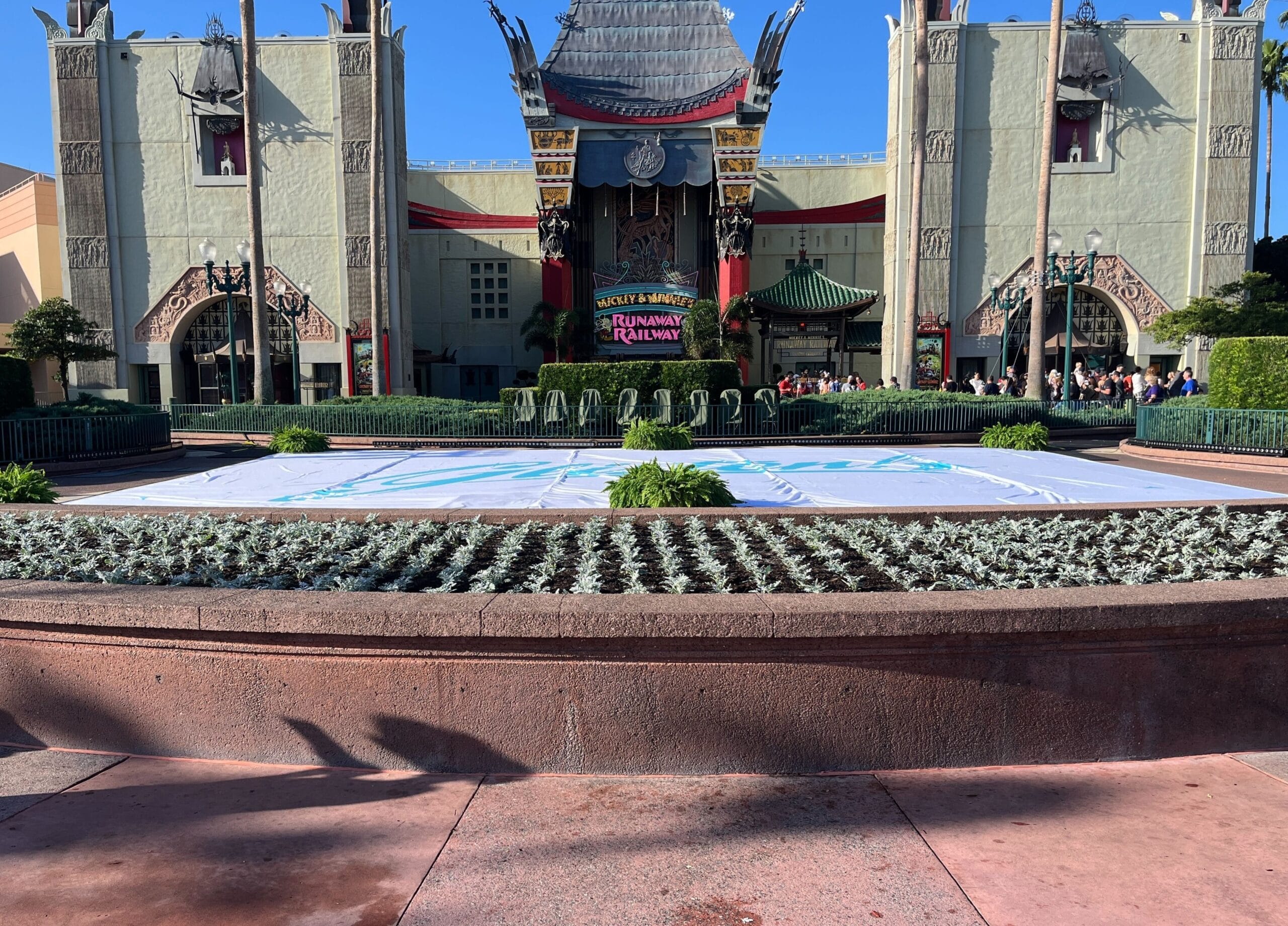 A themed building with ornate details and a Runaway Railway sign, tucked amidst a garden area resembling the charm of Jollywood nights, with low hedges and a circular paved surface in the foreground.