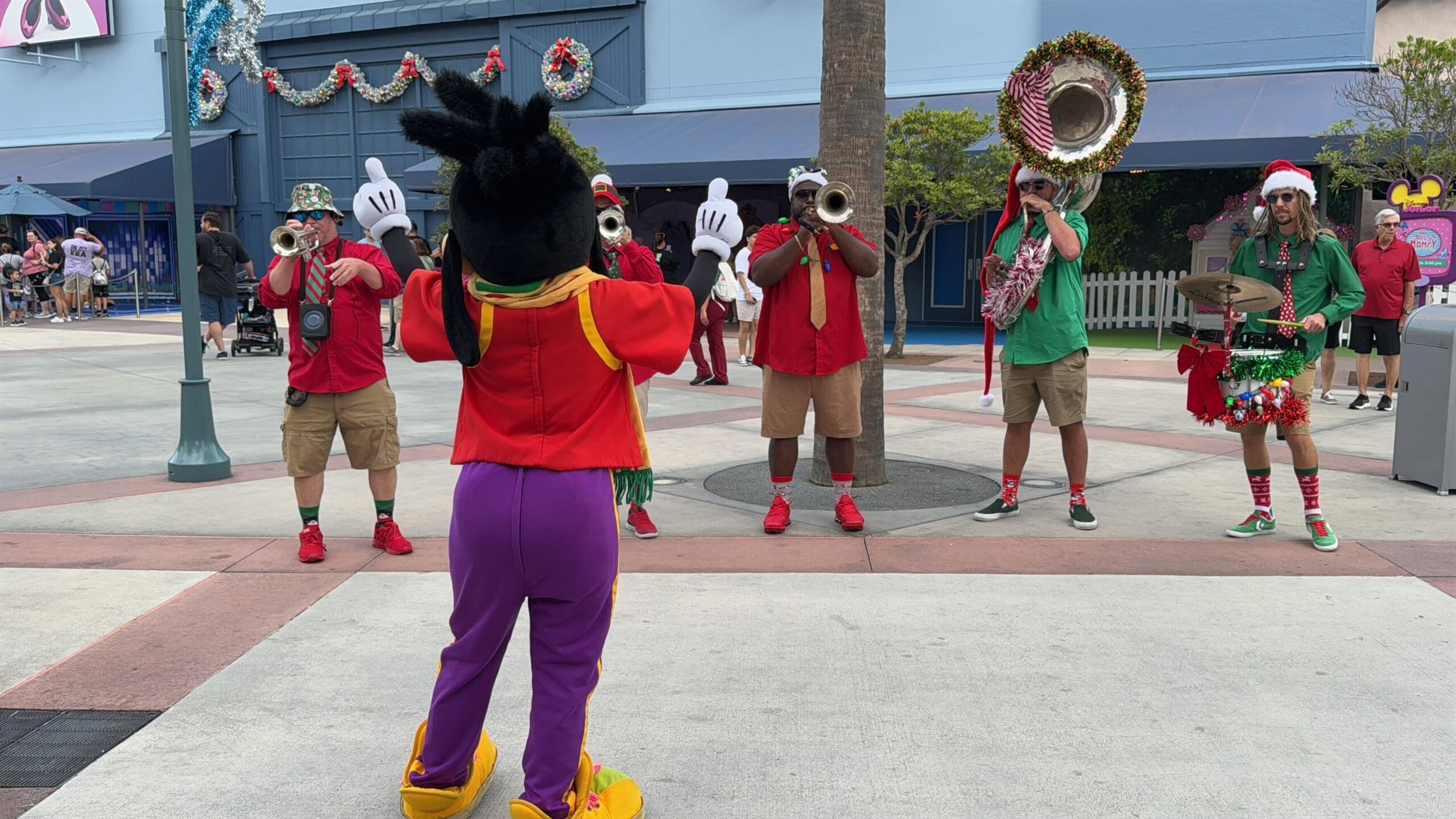 A costumed character with large gloves and colorful outfit directs a brass band playing outdoors; band members wear festive, colorful clothes.