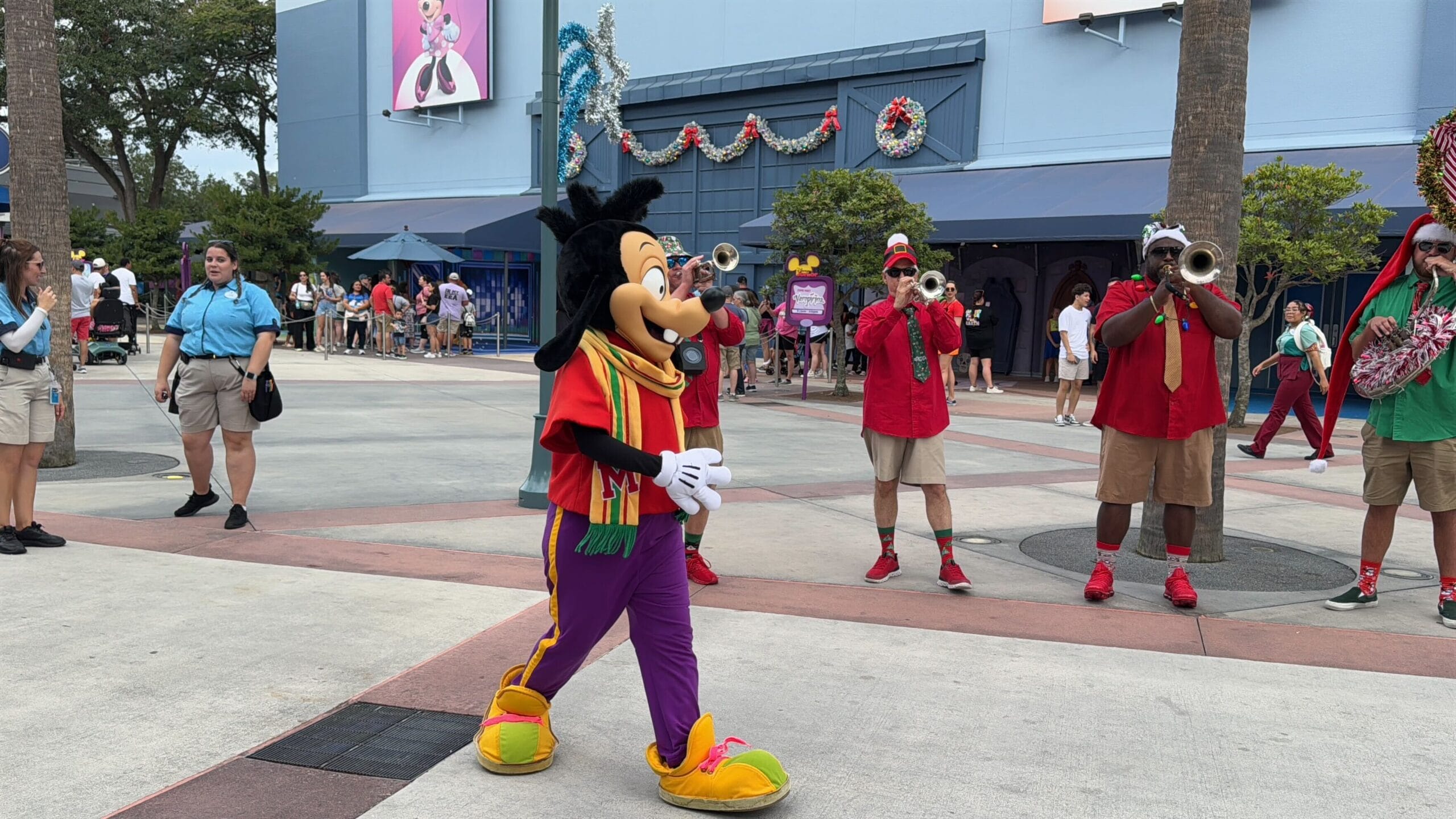 A costumed character walks past musicians in a theme park, with a decorated building in the background.