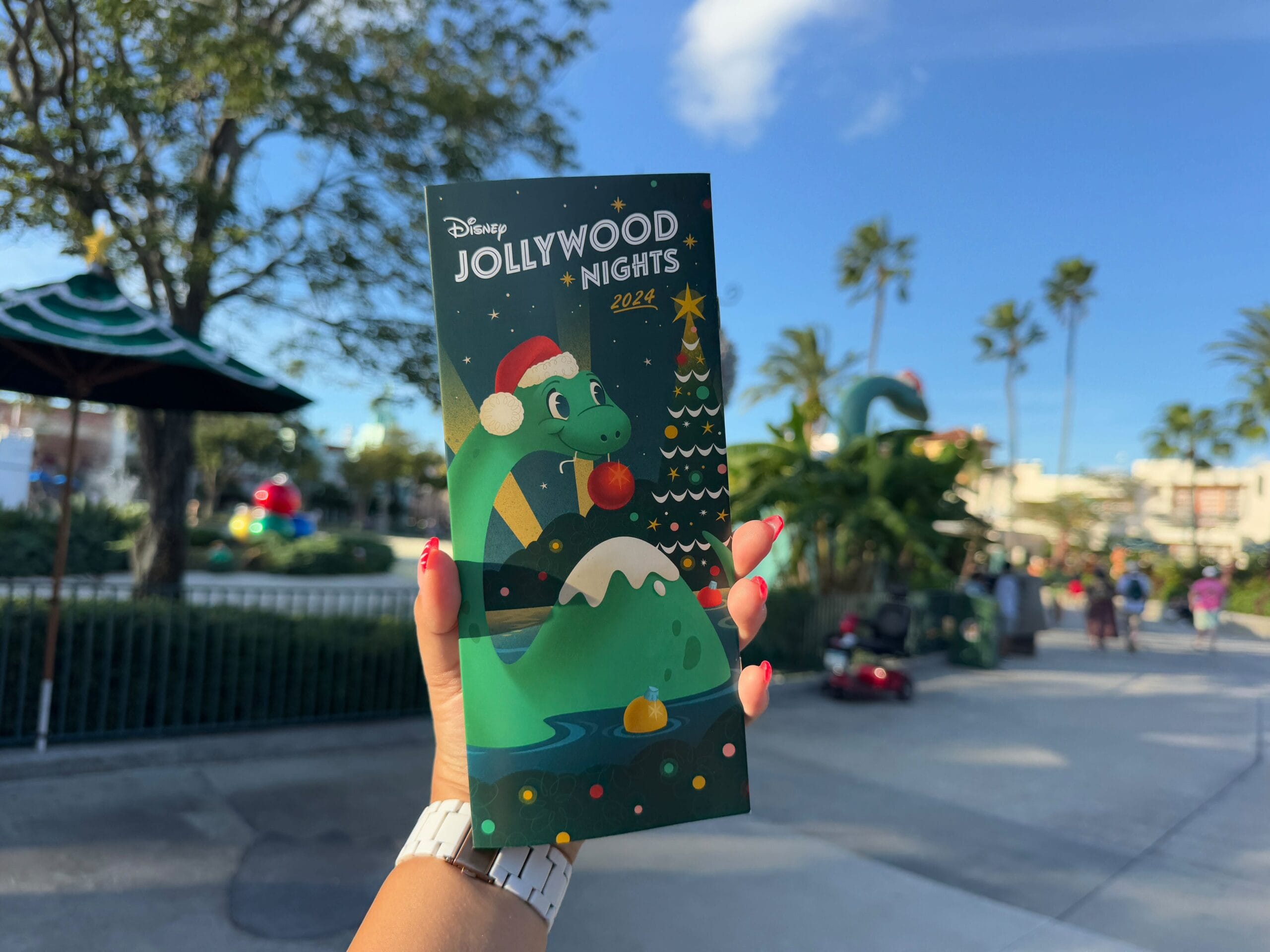 A hand holds a colorful Disney Jollywood Nights 2024 brochure, featuring a dinosaur with a Santa hat and a Christmas tree, against a sunny theme park backdrop with palm trees.