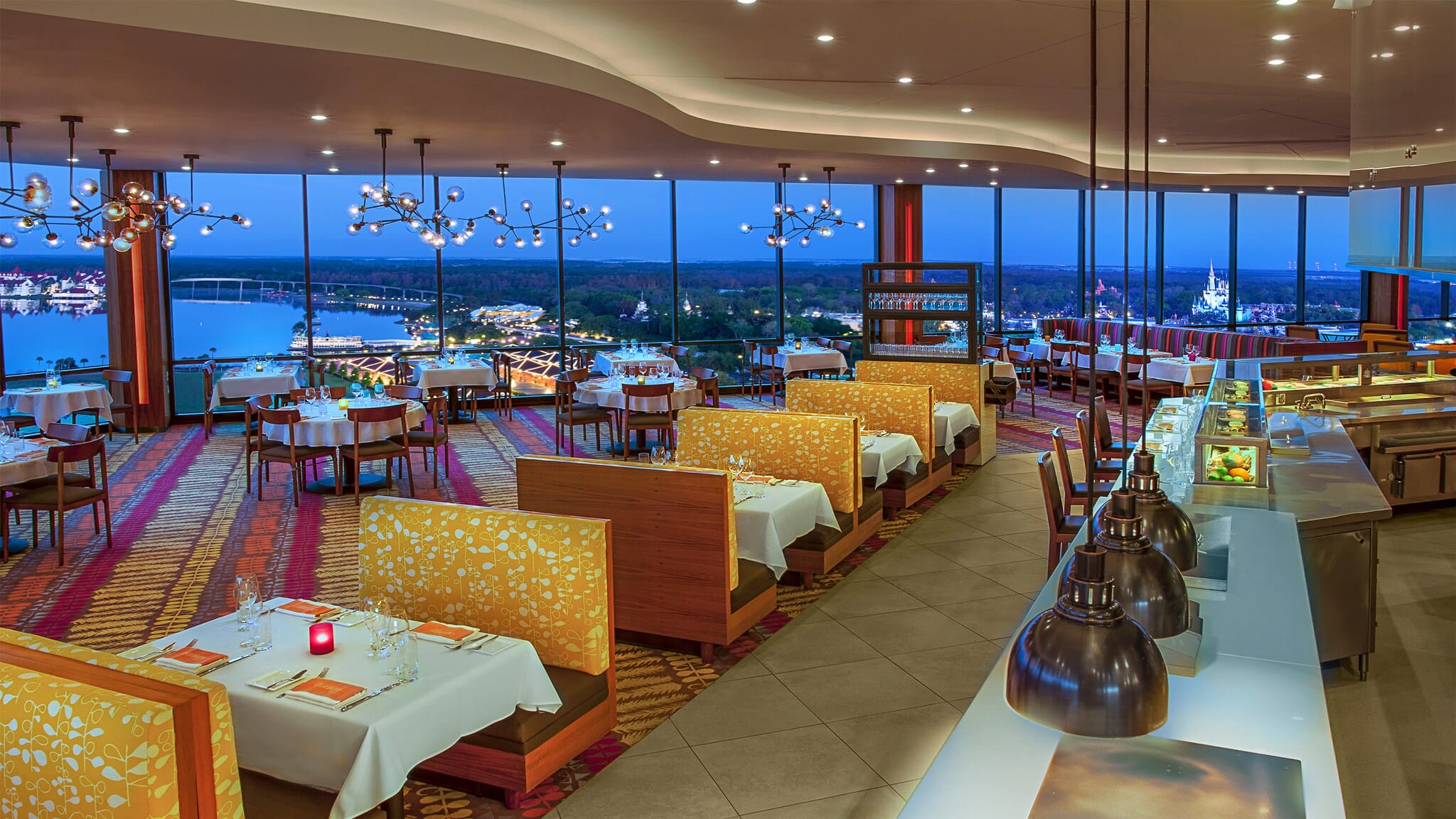 A spacious restaurant with orange decor and wide glass windows offering a panoramic view of a cityscape at dusk. Tables are set for dining, and modern lighting fixtures hang from the ceiling.
